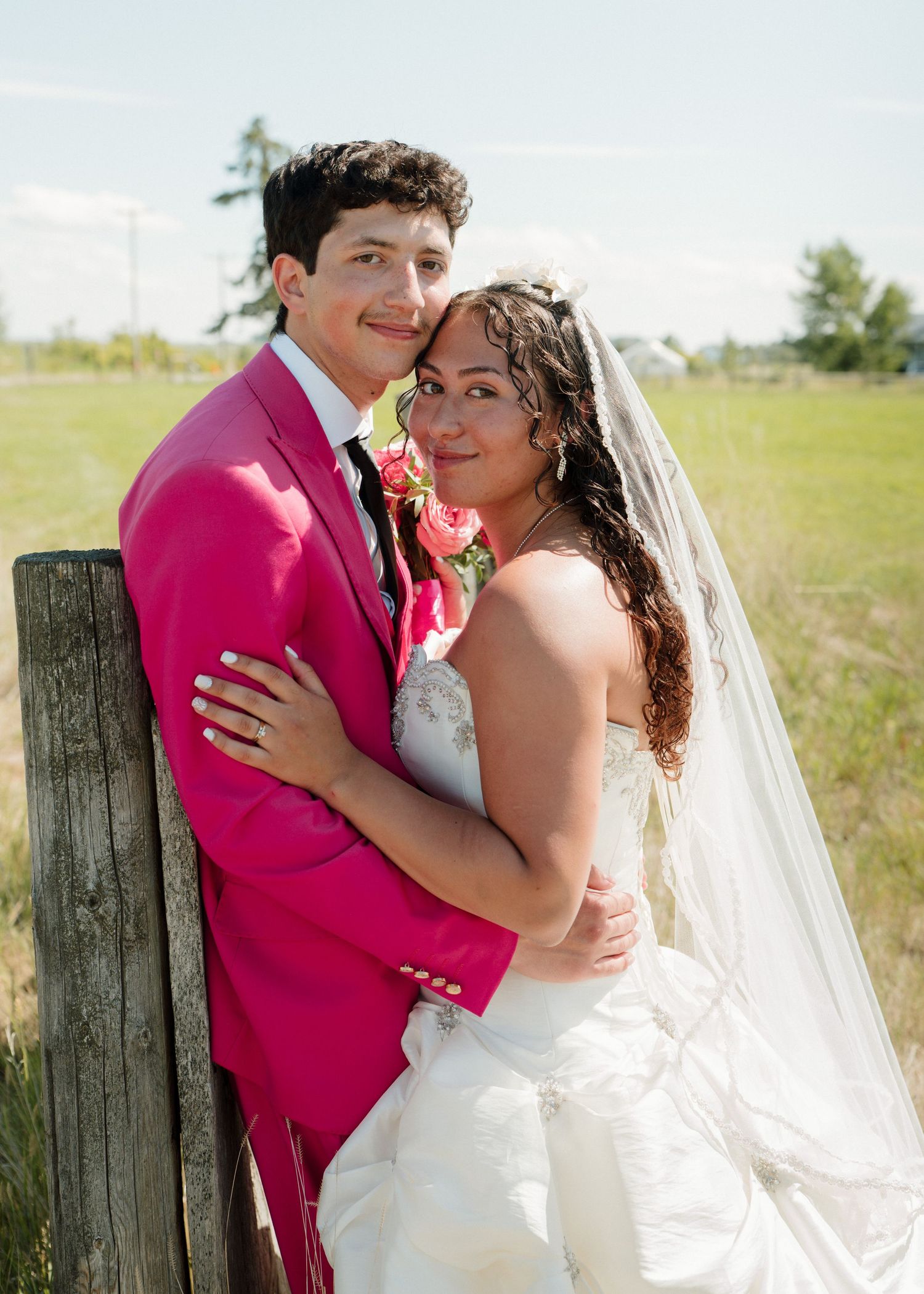 Bride in white gown and veil embraces groom in pink suit by wooden fence in sunny green field.