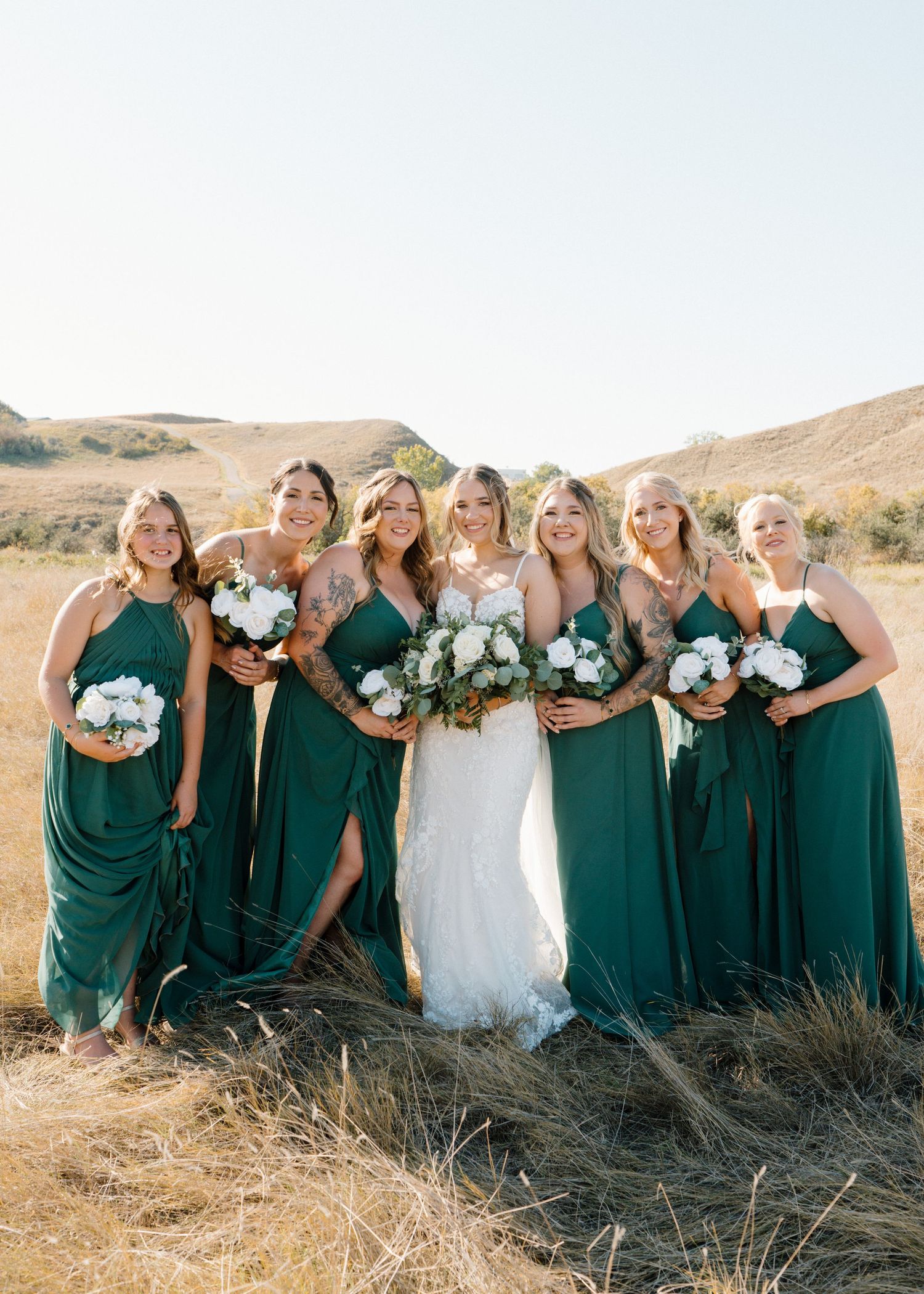 Bride in lace gown with bridesmaids in emerald green dresses holding white bouquets in golden field.