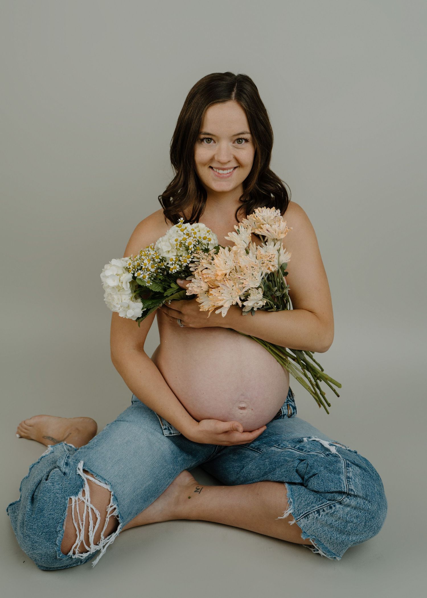 Pregnant woman sits cross-legged holding a bouquet of peach and white flowers in a studio.