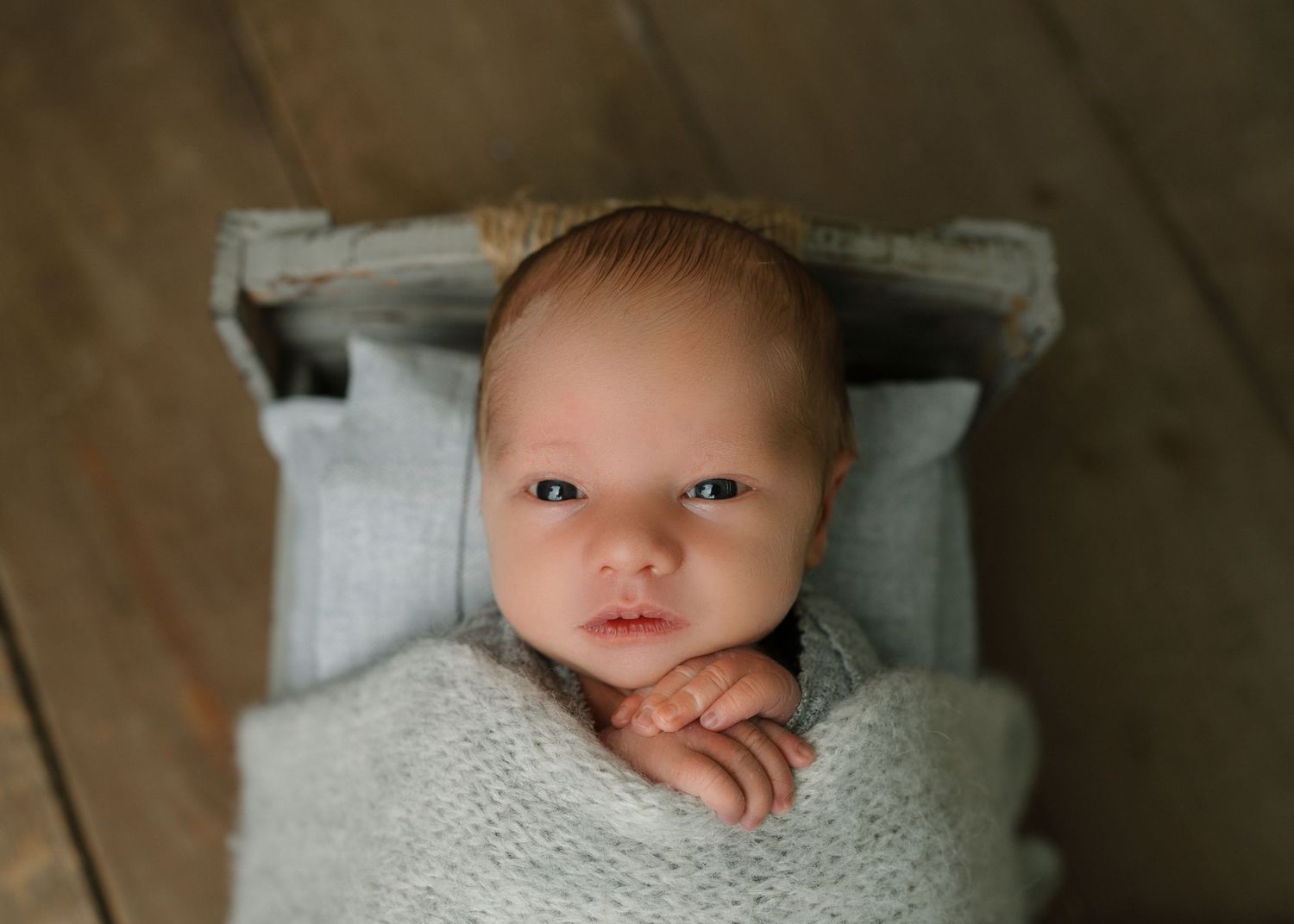 Newborn baby with eyes open wrapped in a soft gray knit blanket lying in a rustic wooden mini bed.