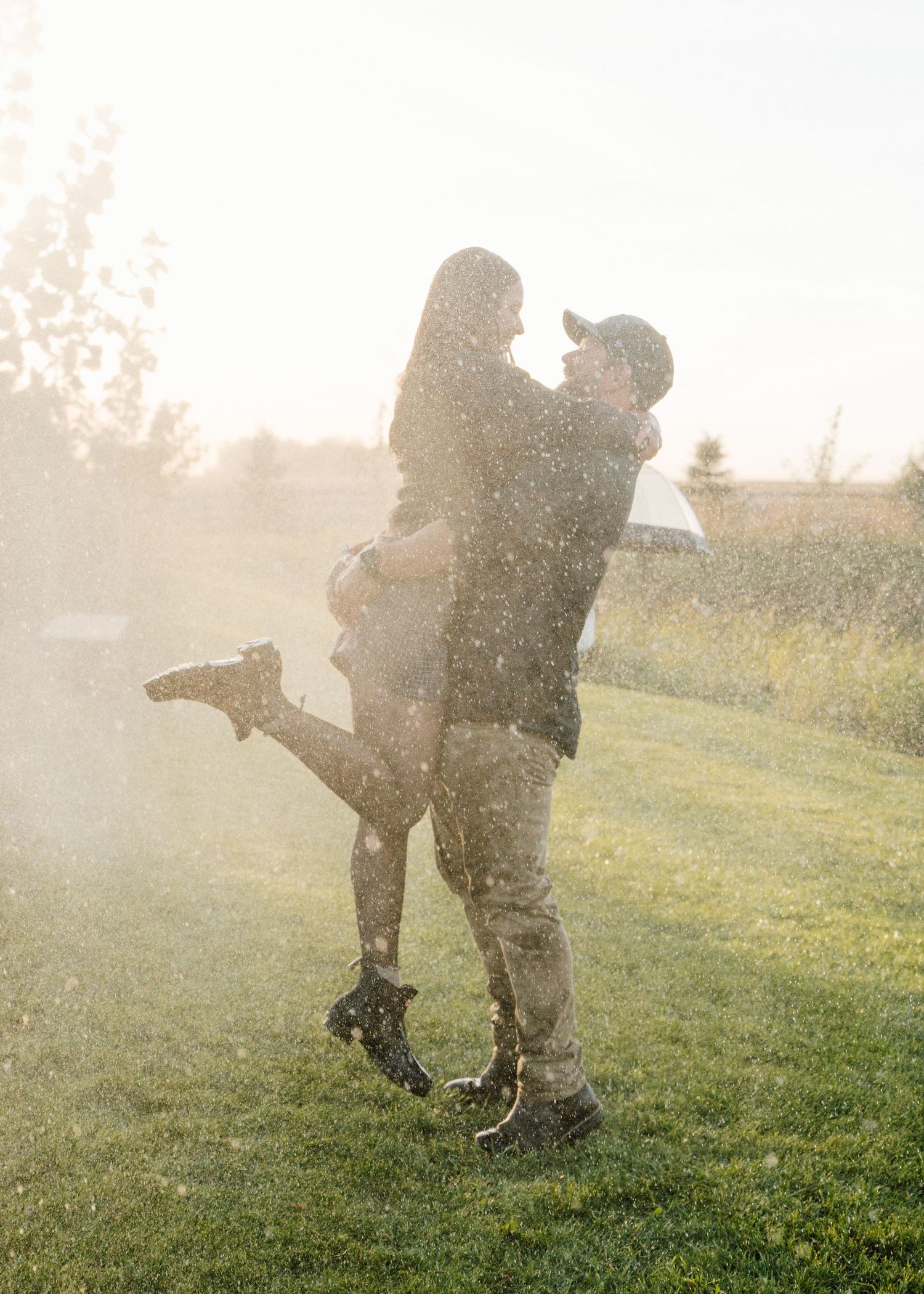 Couple embracing in golden sunlight rain on green grass, romantic outdoor moment with warm backlit glow.