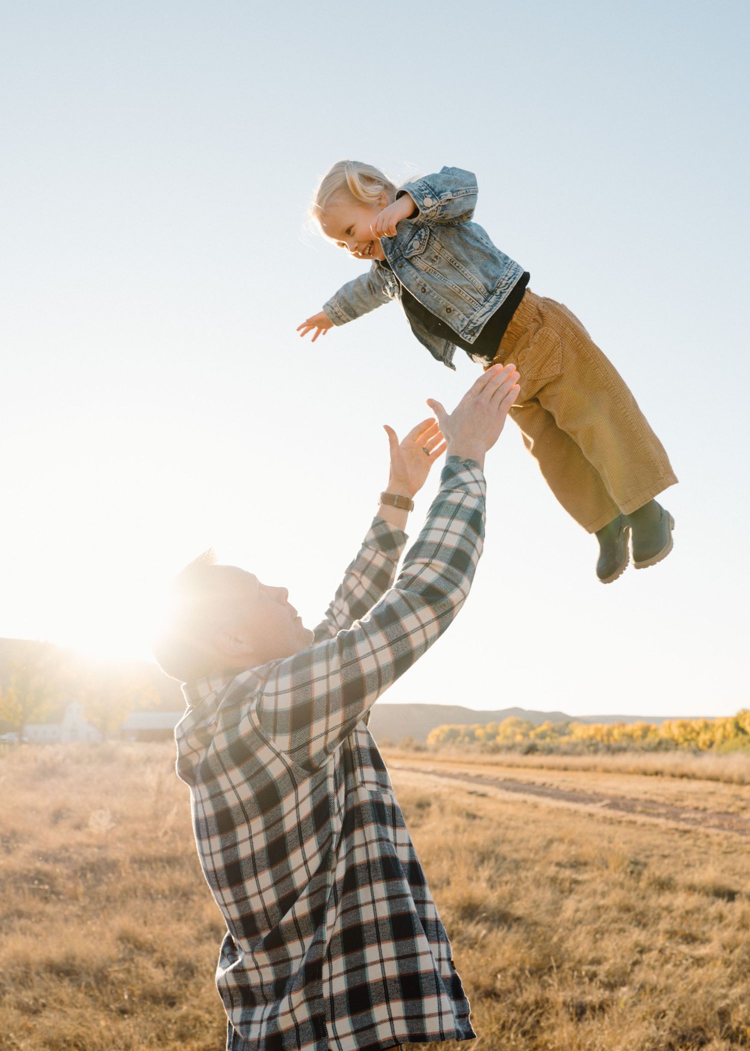 Father tosses laughing toddler high into the air in a sunny open field at golden hour.
