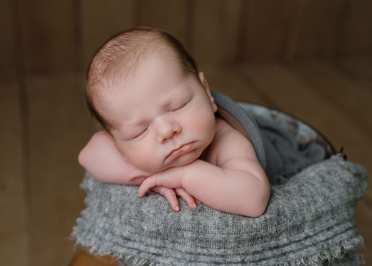 Peaceful sleeping newborn baby posed in gray knit bucket prop on wooden floor.