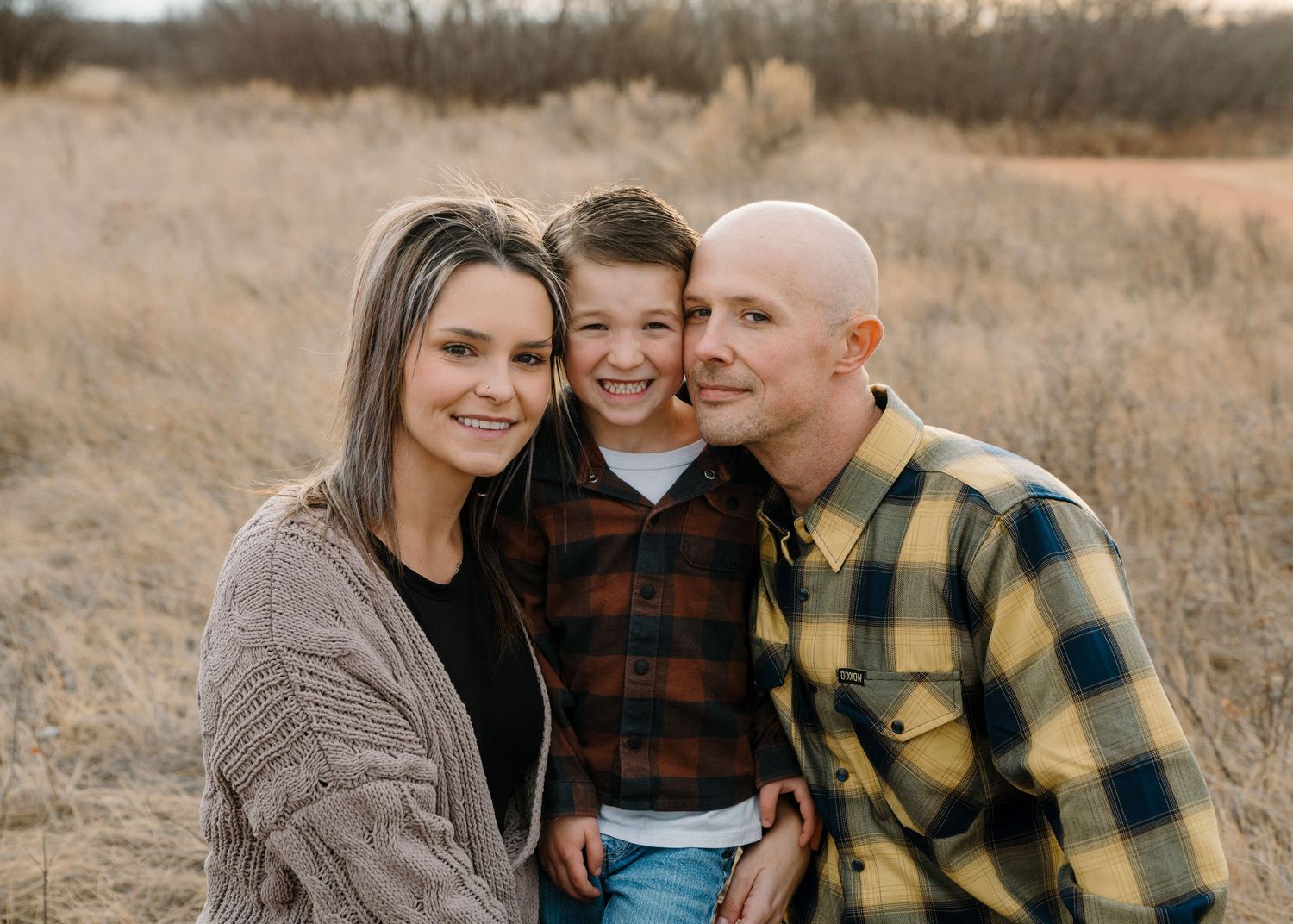 Happy family of three posing outdoors in a golden field, parents smiling with young boy between them.