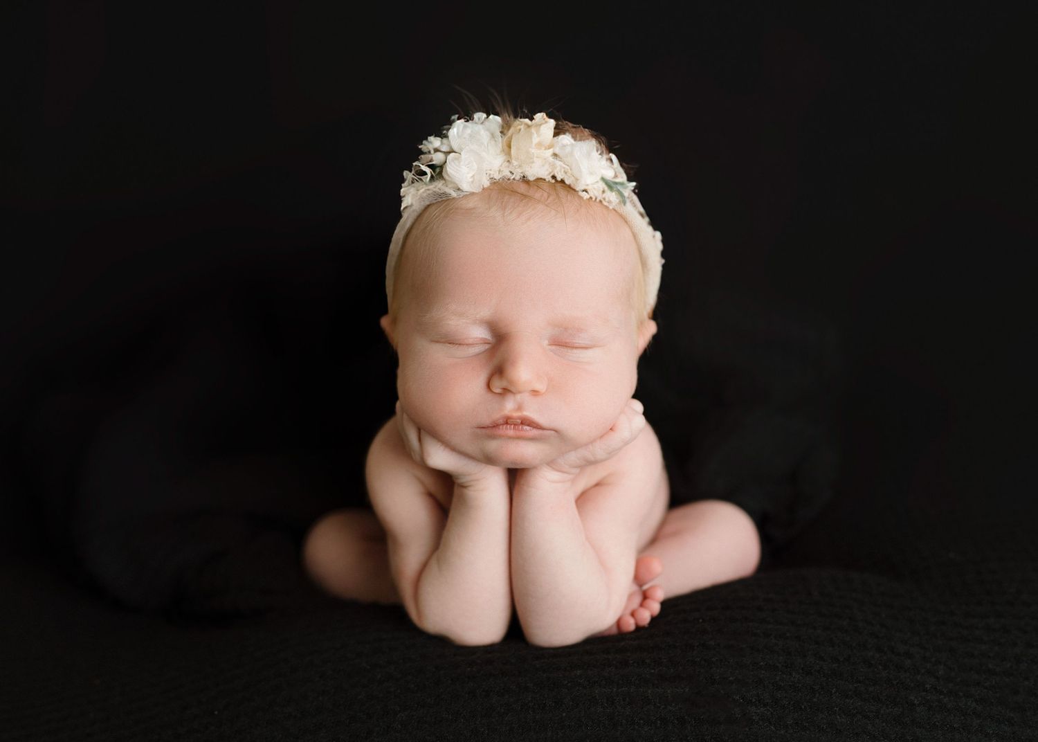 Peaceful newborn baby sleeping in froggy pose wearing delicate floral headband on dark background.