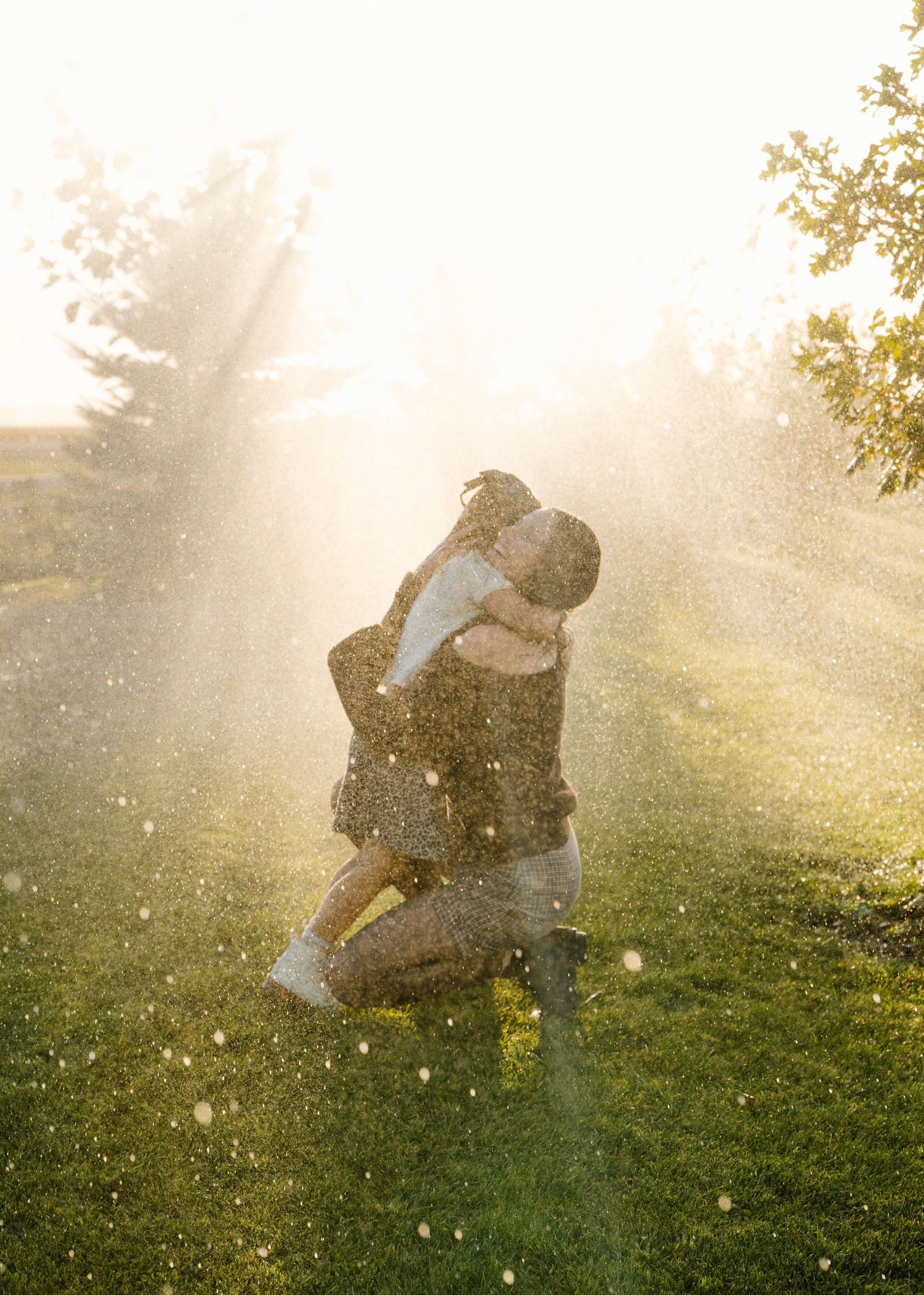 Couple embracing on grass in golden sunlight with water droplets sparkling around them in a romantic outdoor setting.