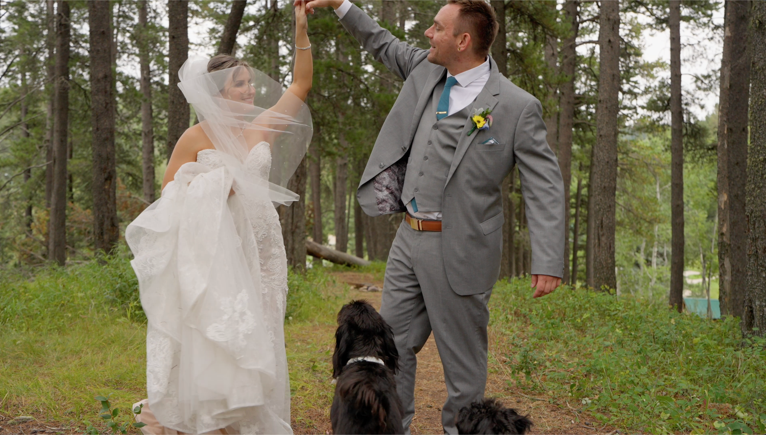 Bride and groom dancing in forest with two black dogs, groom in gray suit twirling bride in white wedding dress.