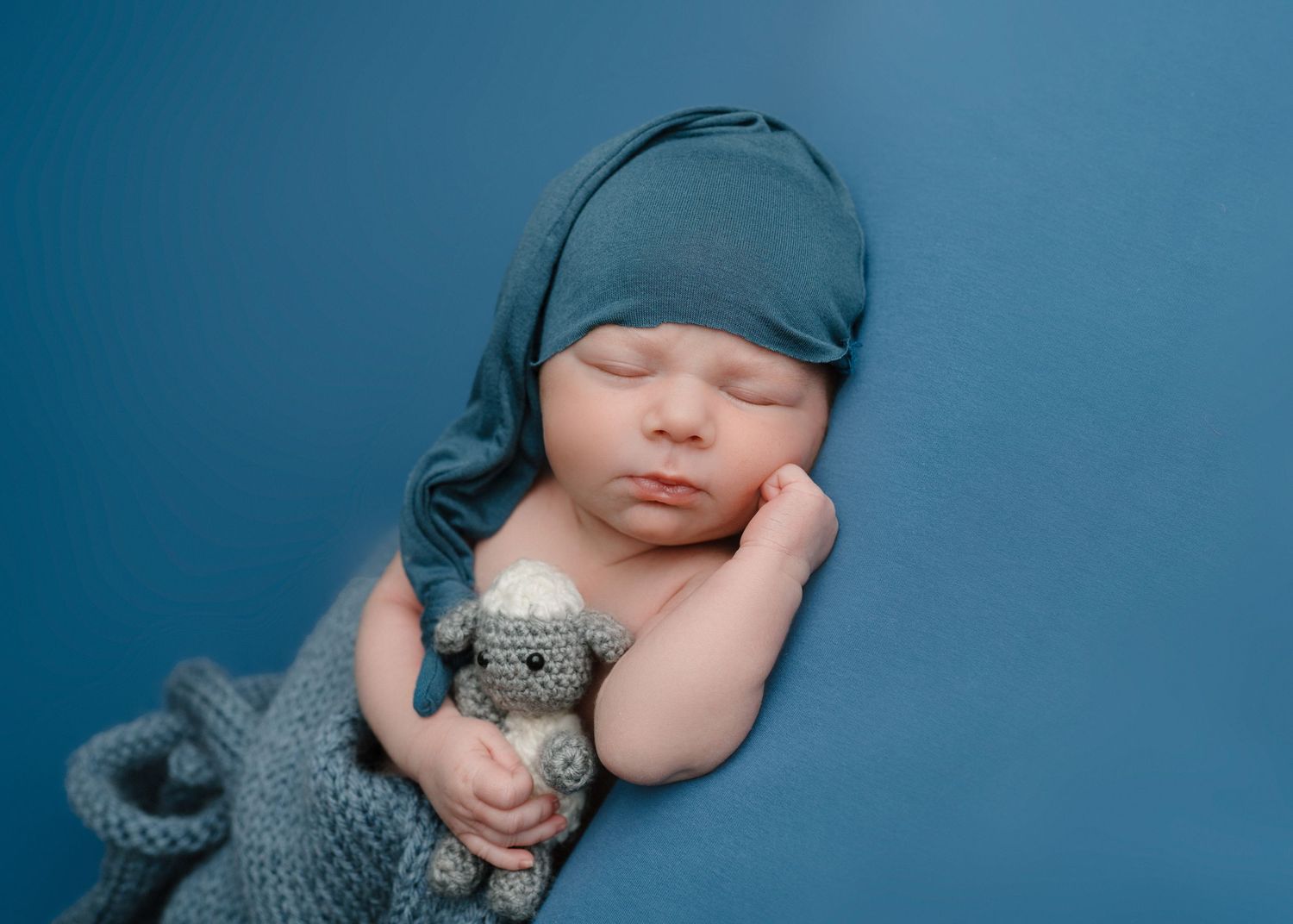Peaceful newborn baby in teal sleepy hat holding a small crocheted toy on a blue backdrop.