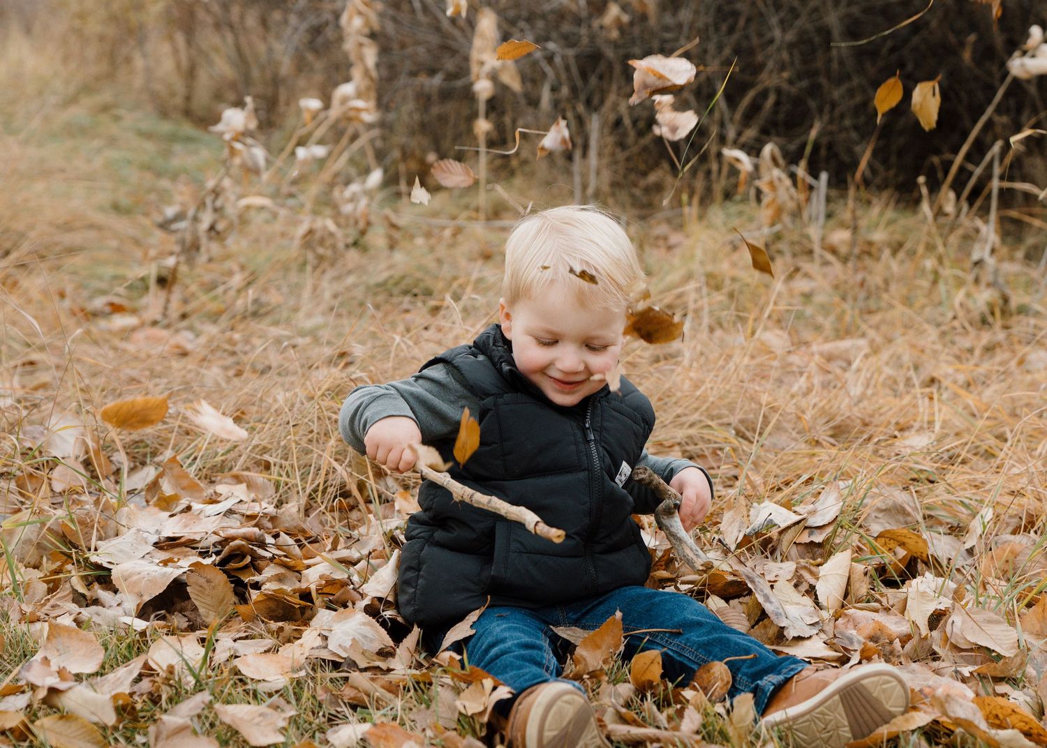 Cheerful toddler sitting in fallen autumn leaves playing with a stick in a golden field.
