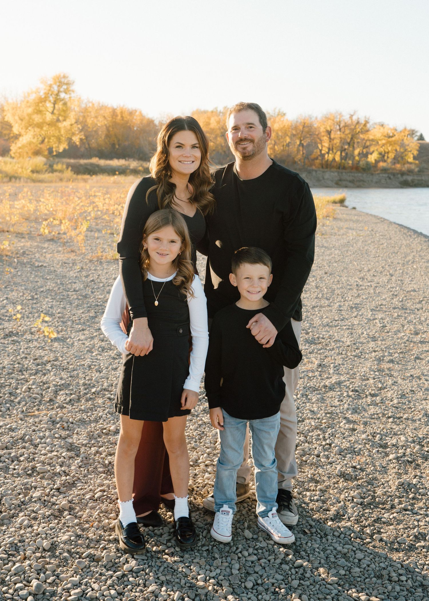 Family of four poses together on a rocky shoreline surrounded by golden autumn trees at sunset.