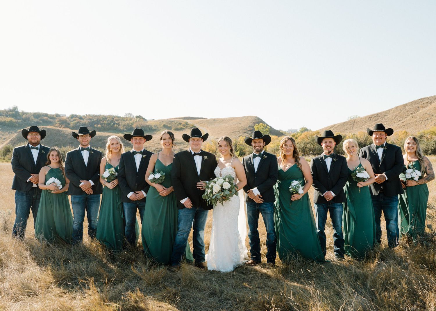 Western wedding party in golden hills, groomsmen in cowboy hats, bridesmaids in emerald green gowns.