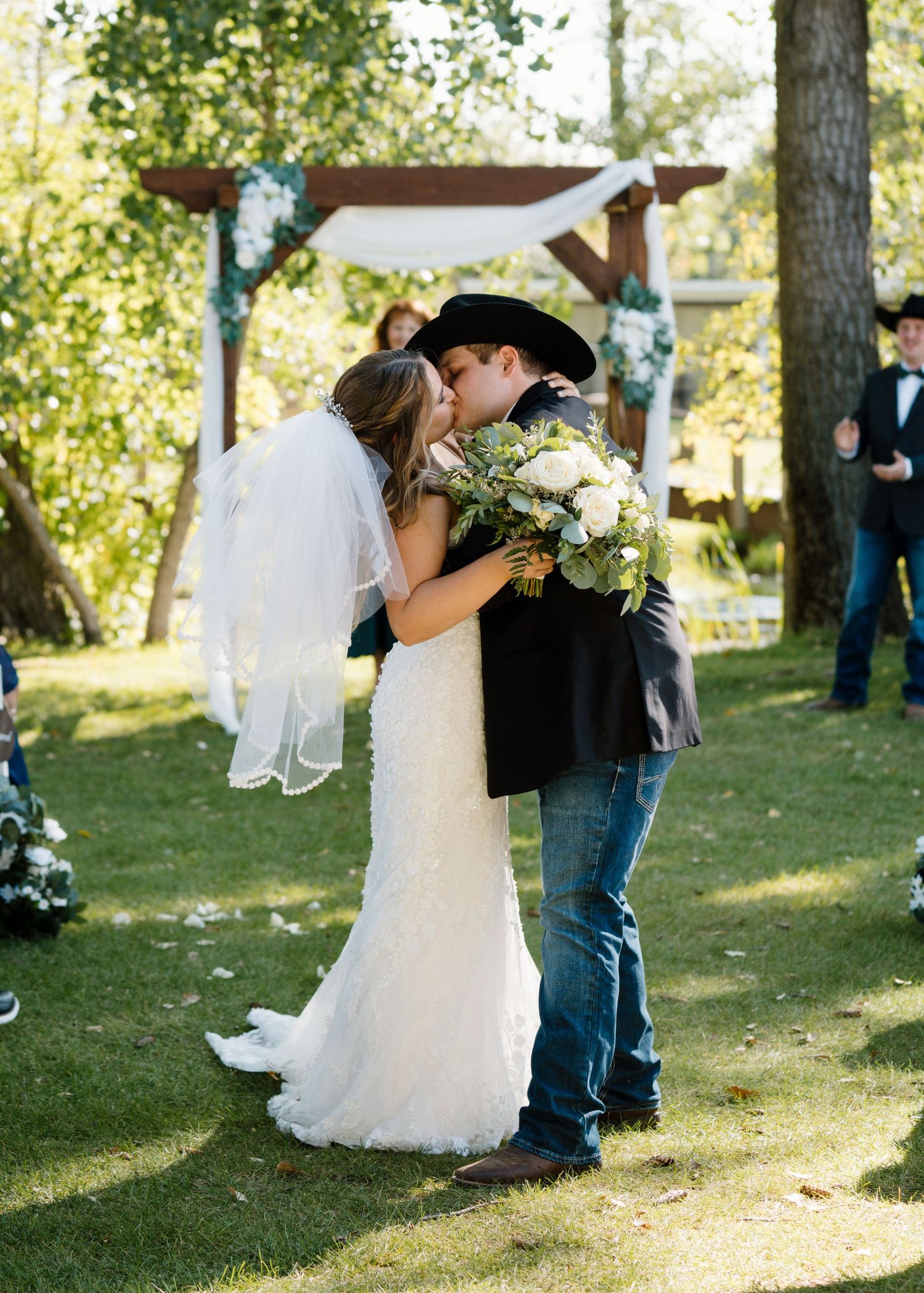 Bride and groom share first kiss at outdoor wedding ceremony under rustic wooden arch with floral decorations.