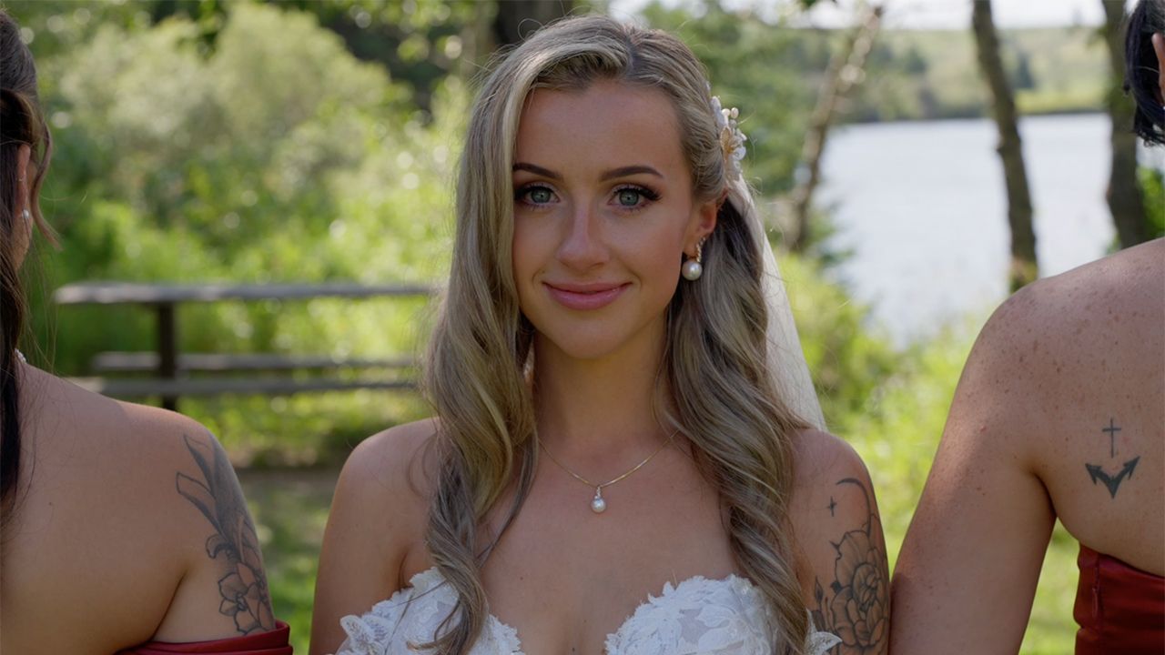 Smiling blonde bride in white strapless wedding gown with floral hair accessory standing outdoors near a lake.
