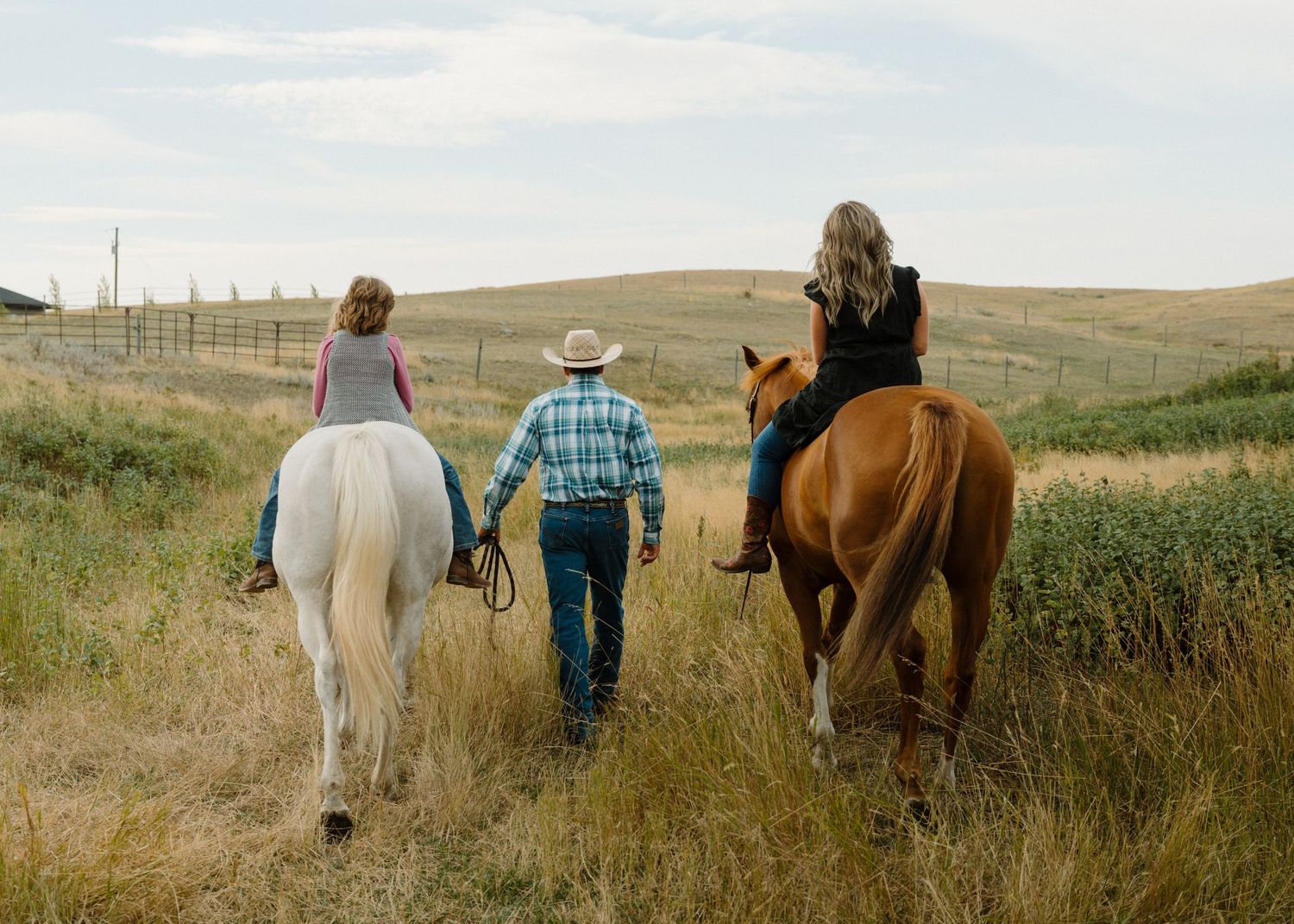 Family walking with two horses through a golden grass field, man in plaid shirt holding hands with riders.