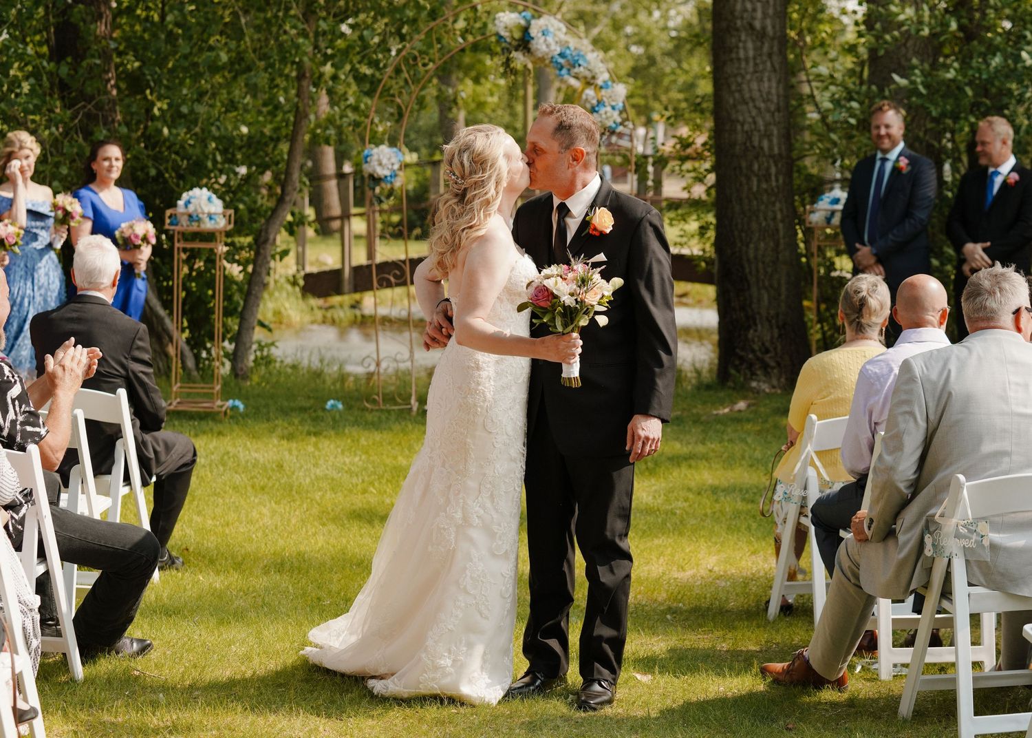 Bride and groom share first kiss at outdoor garden wedding ceremony with seated guests.
