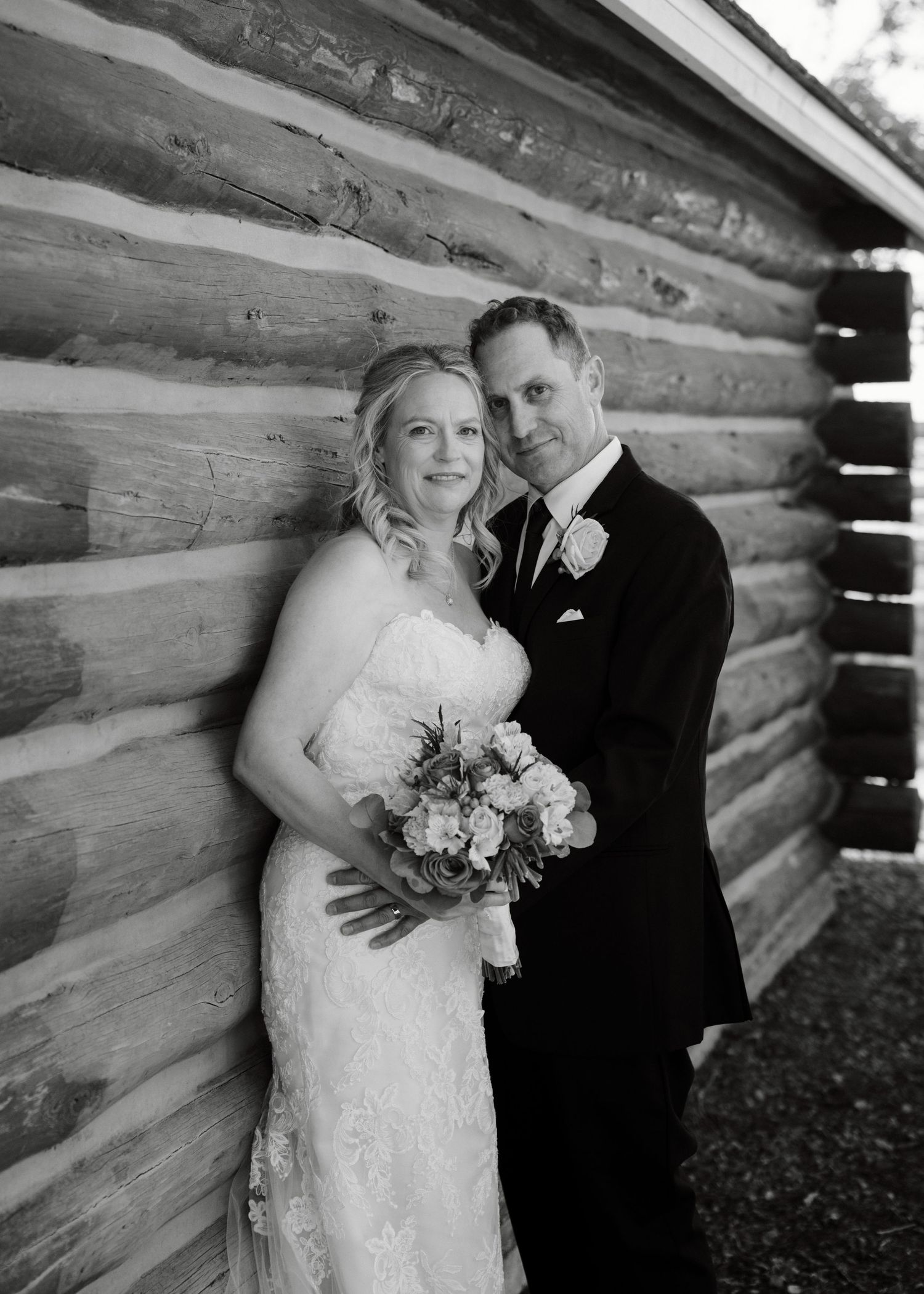 Black and white portrait of bride and groom embracing against a rustic log cabin wall.