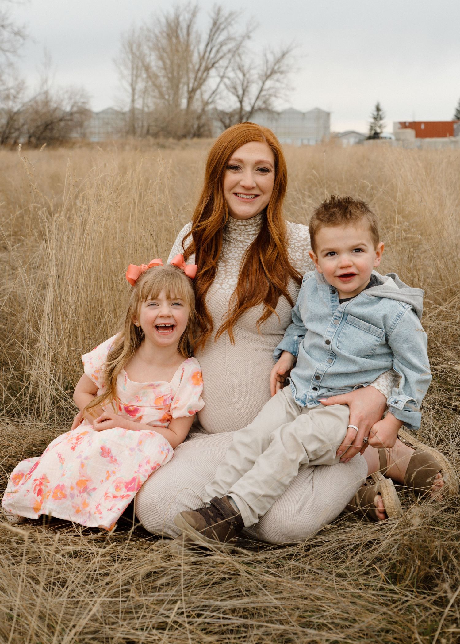 Pregnant redheaded mother sitting in golden grass field with two young children smiling.