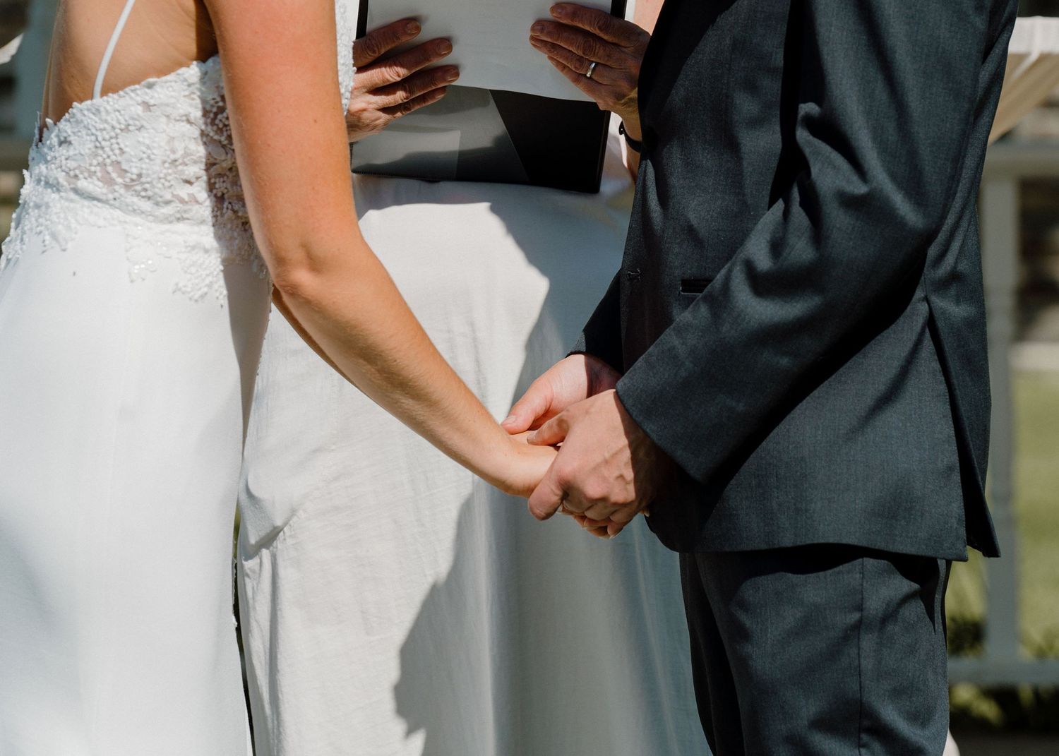 A bride and groom hold hands during their wedding ceremony, she in a white lace gown, he in a gray suit.