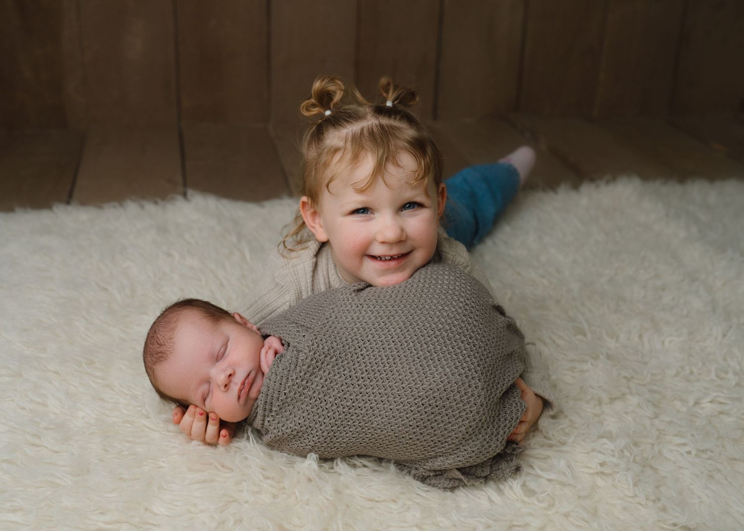Smiling toddler girl with pigtails lying on fluffy white rug beside sleeping swaddled newborn sibling.