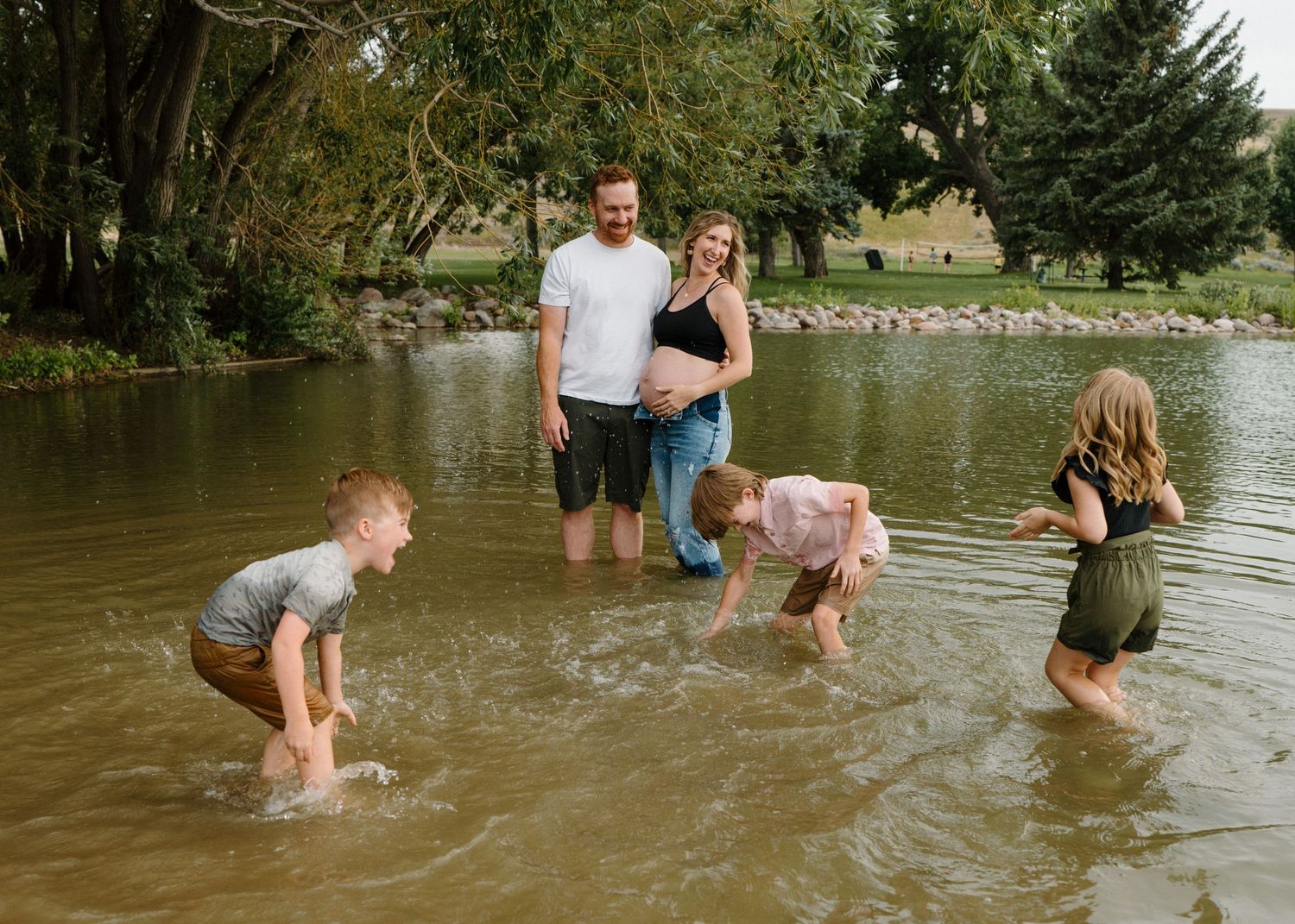 Family with pregnant mother playing in a shallow pond surrounded by lush green trees.