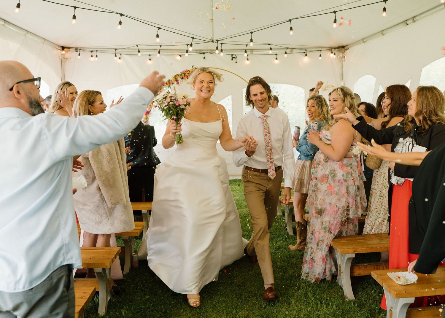Bride and groom joyfully walking through cheering guests under a tent at their wedding reception.