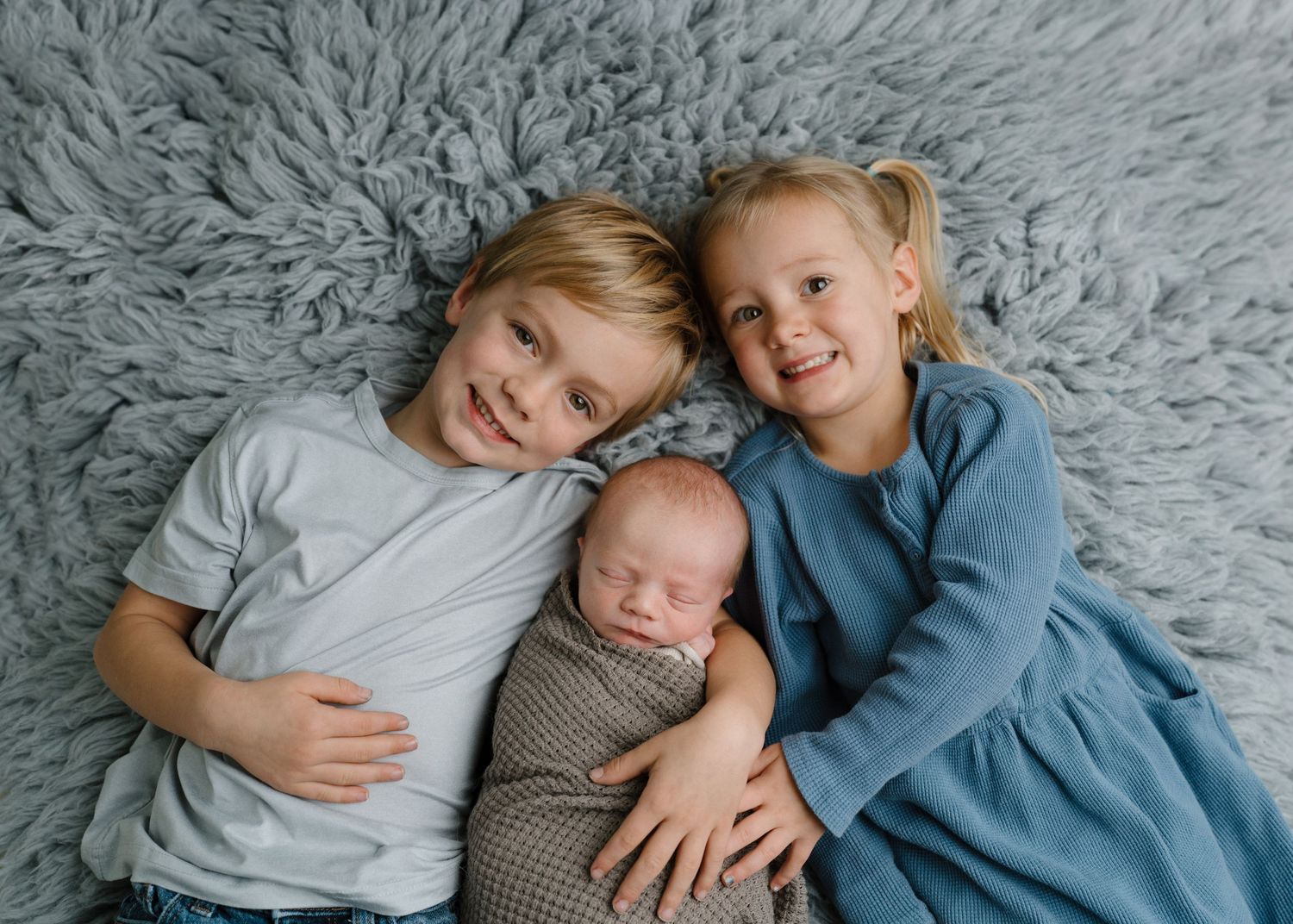 Two blonde siblings lying on grey fur rug smiling while gently holding sleeping swaddled newborn.
