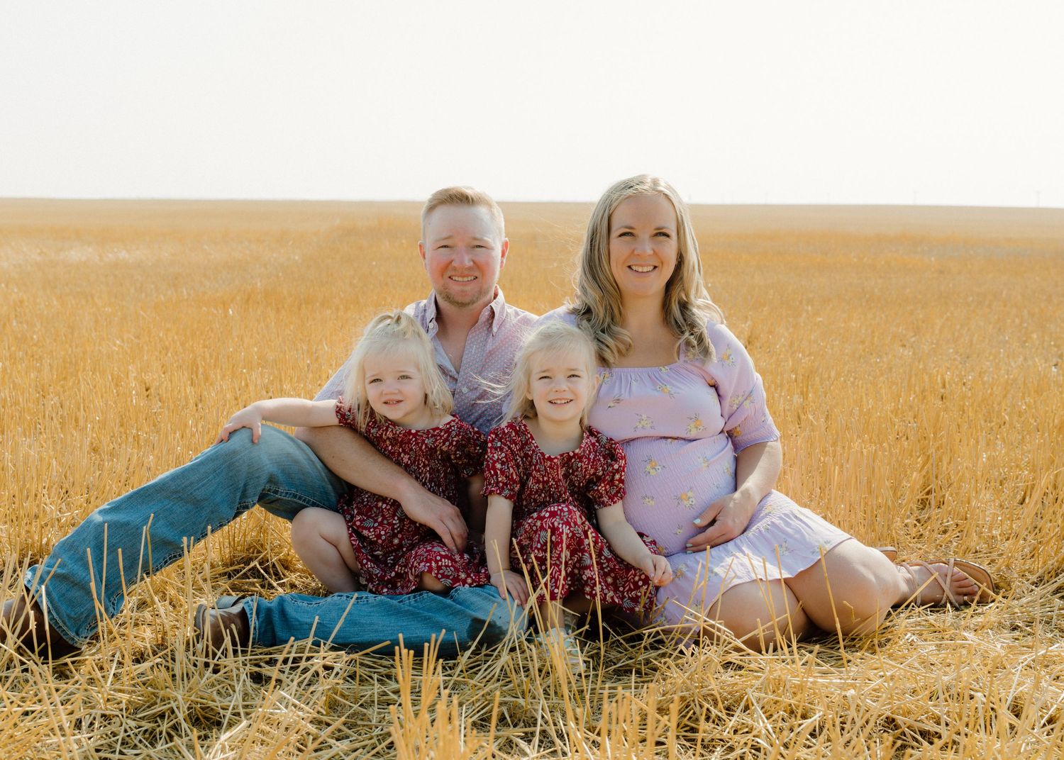 Happy family maternity portrait sitting in golden wheat field with two blonde girls in red dresses.