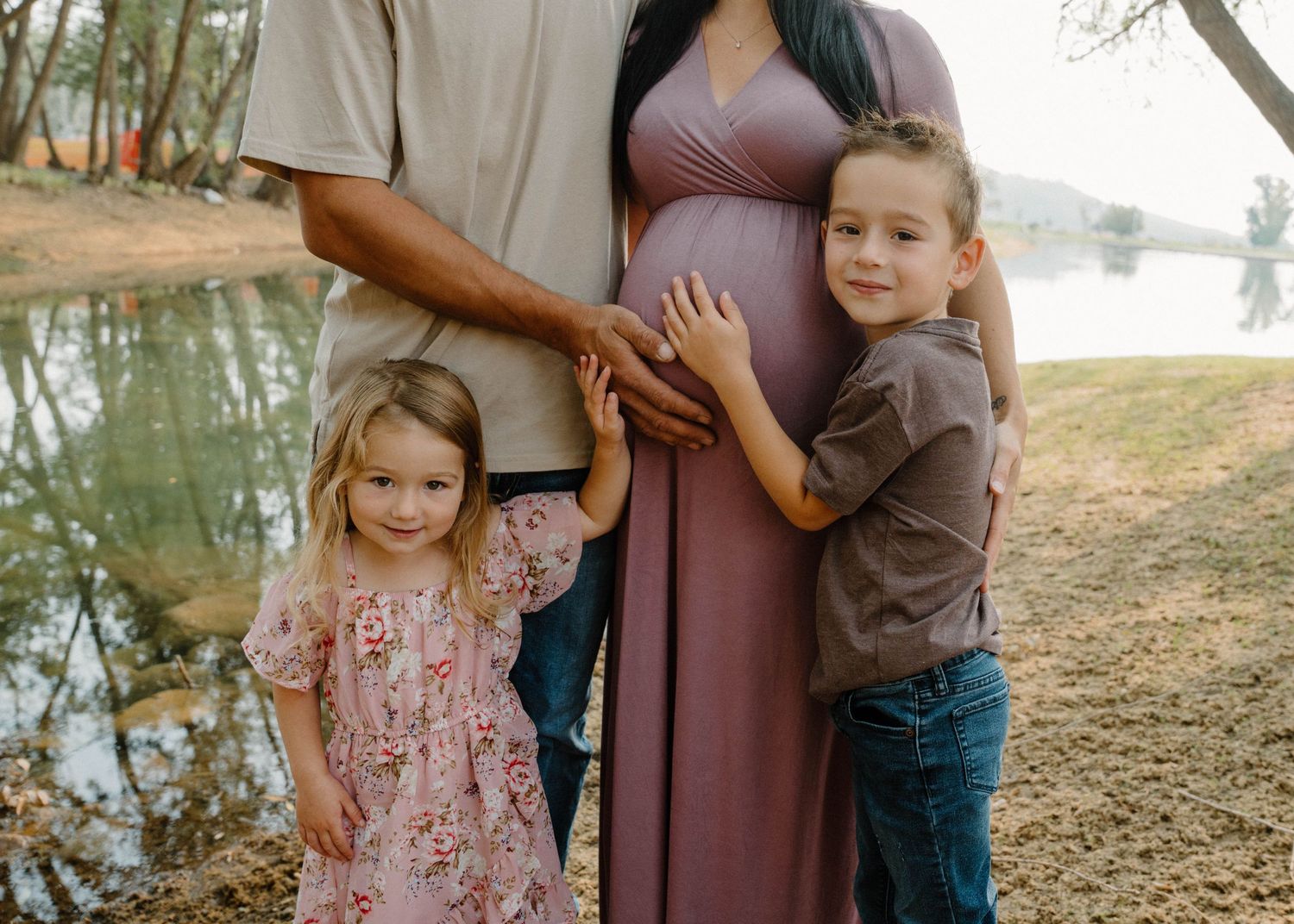 Two young children hugging pregnant mothers baby bump outdoors near lake during family maternity photo session.
