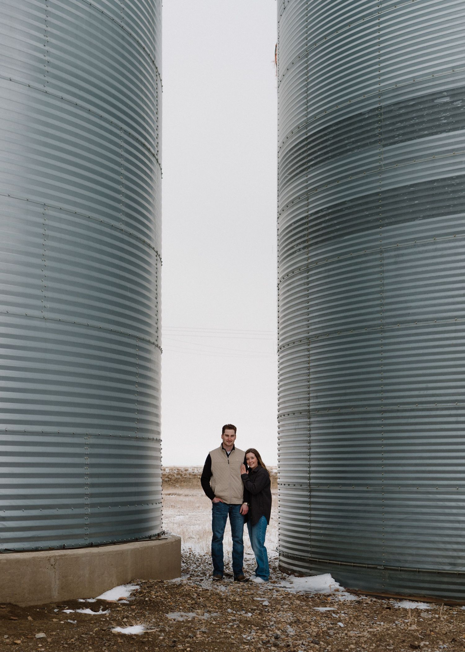 Engaged couple standing between large silver grain silos on a snowy farm in winter.