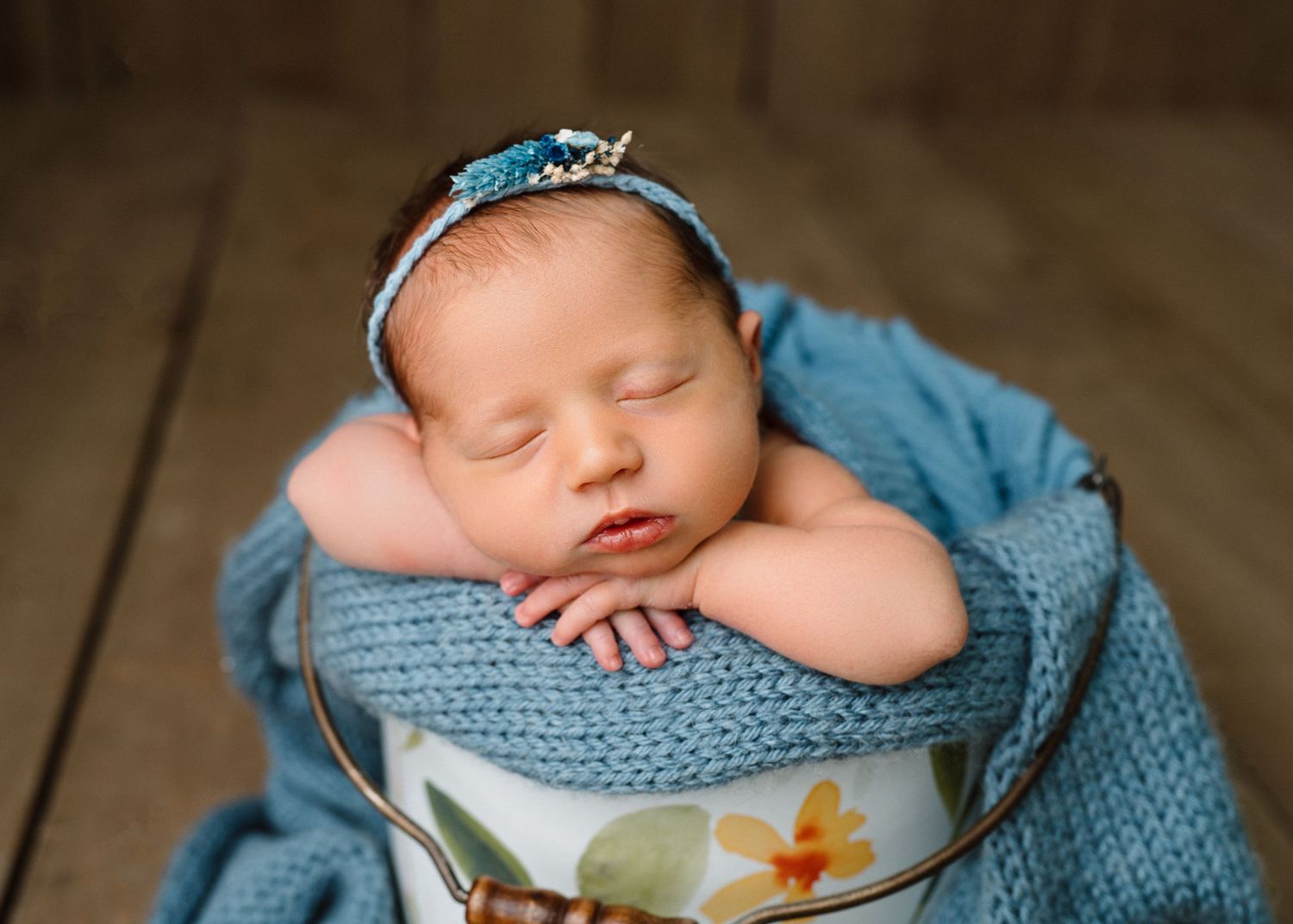 Sleeping newborn baby girl in a floral bucket wrapped in teal knit blanket wearing a delicate headband.