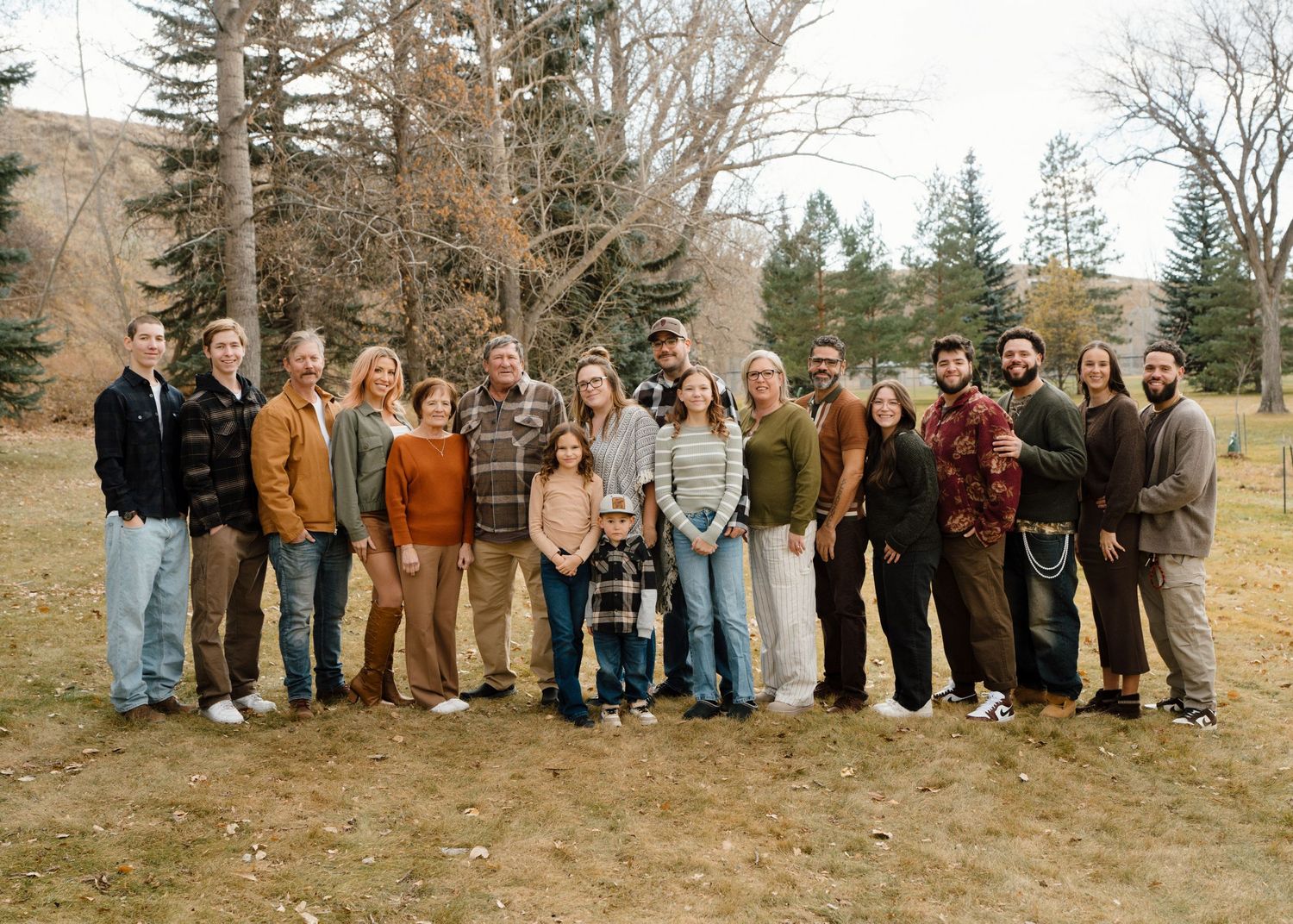 Large extended family group of twenty posing outdoors in coordinated earth-tone outfits in fall setting.