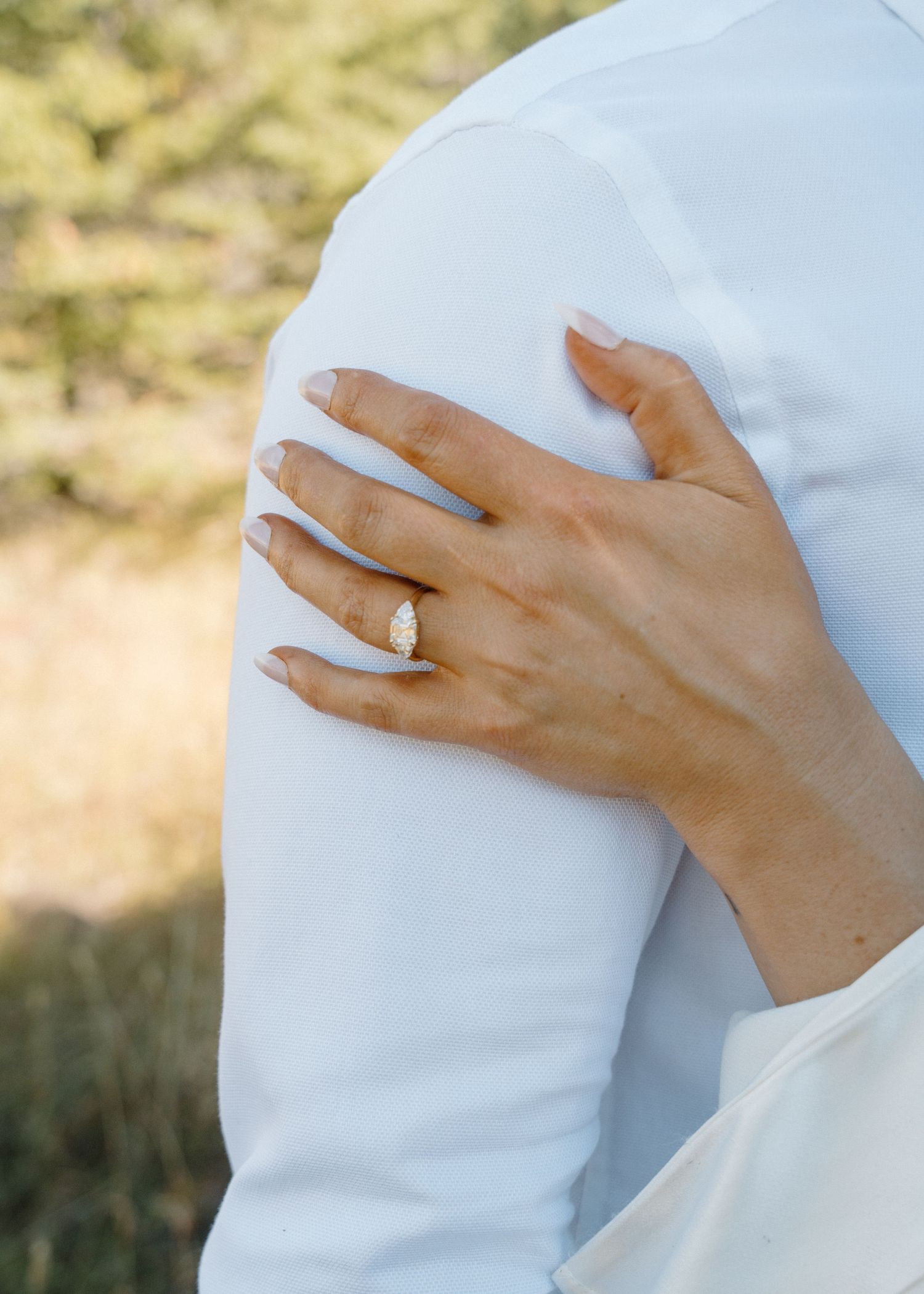 Close-up of bride's hand with diamond ring resting on groom's white dress shirt shoulder.