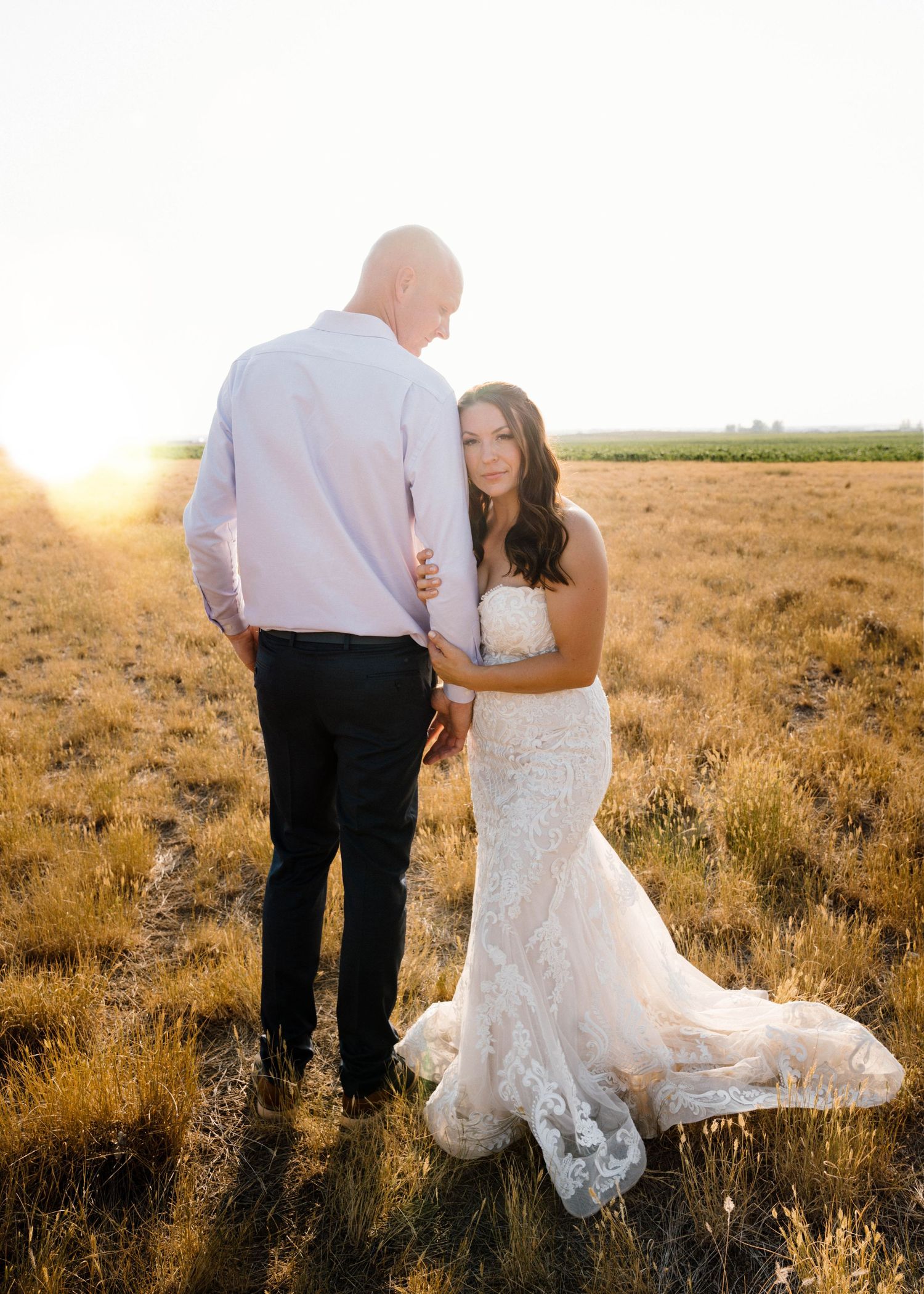 Bride in lace gown leans against groom as golden sunset glows behind them in an open field.