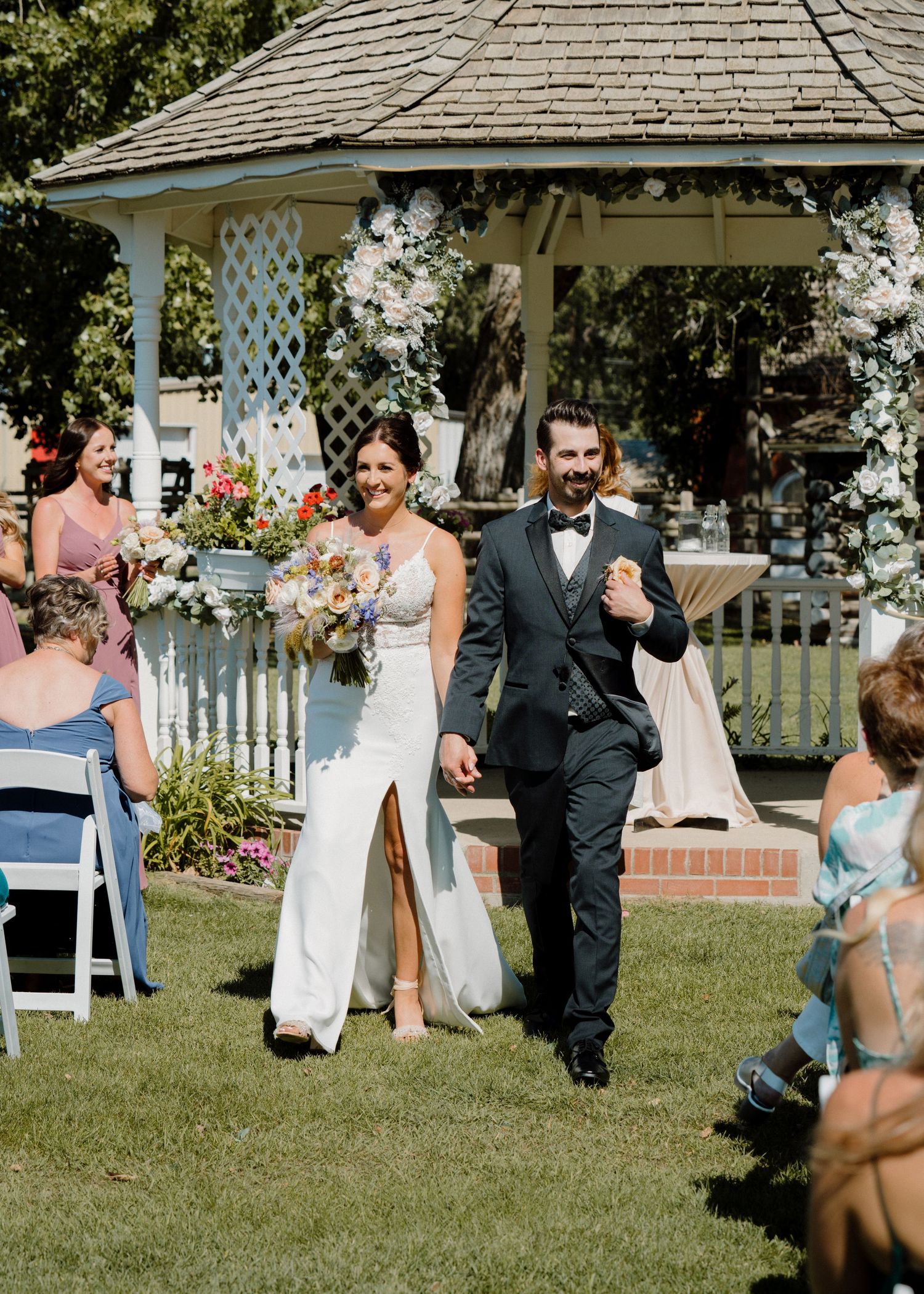 Newlyweds walk hand in hand down the aisle past a floral gazebo at an outdoor garden wedding.