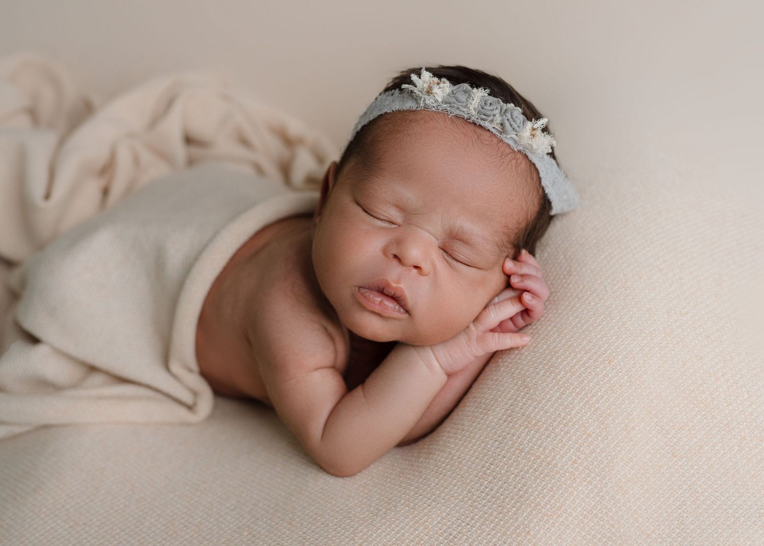 Sleeping newborn baby girl wearing a delicate floral headband wrapped in a cream blanket posed on beige backdrop.