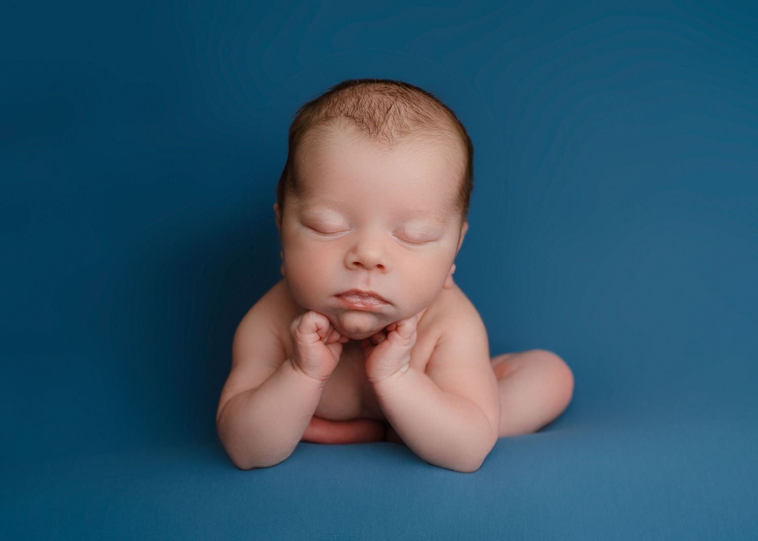 Newborn baby posed in froggy position with hands under chin sleeping on a blue photography backdrop.