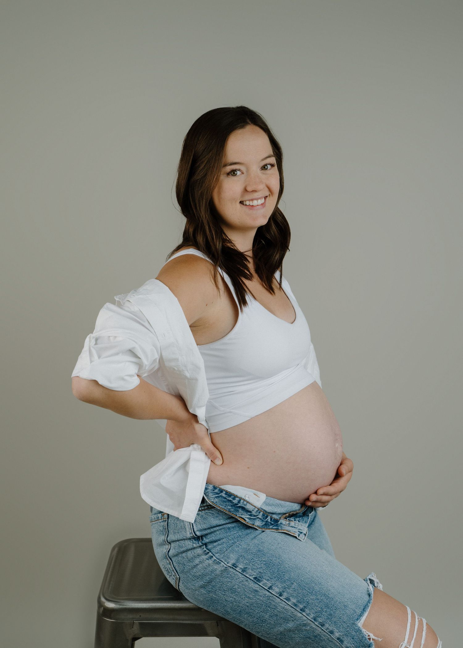 Pregnant woman in white tank and ripped jeans sits on stool showing baby bump in studio.