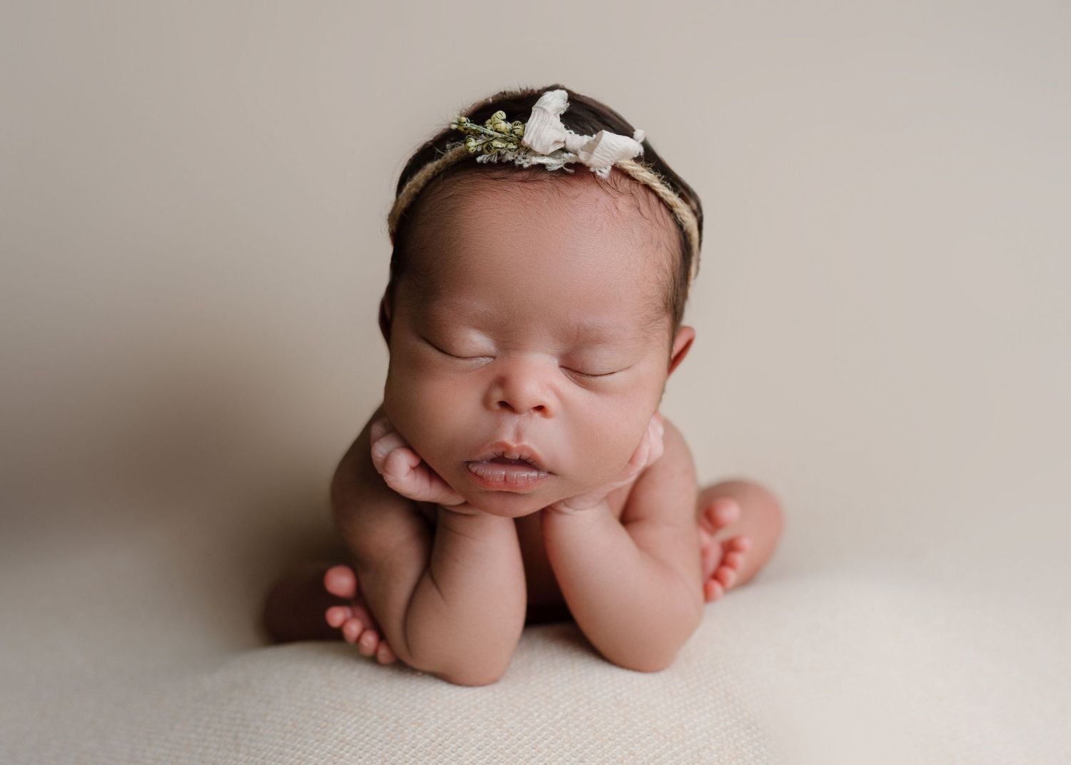 Sleeping newborn baby posed in froggy position wearing a delicate floral headband on cream backdrop.