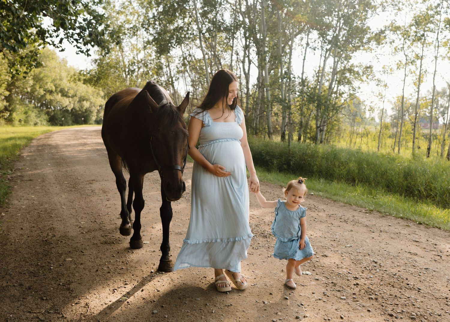 Pregnant woman in a light blue dress walking with a toddler girl and a dark horse on a sunny dirt path.
