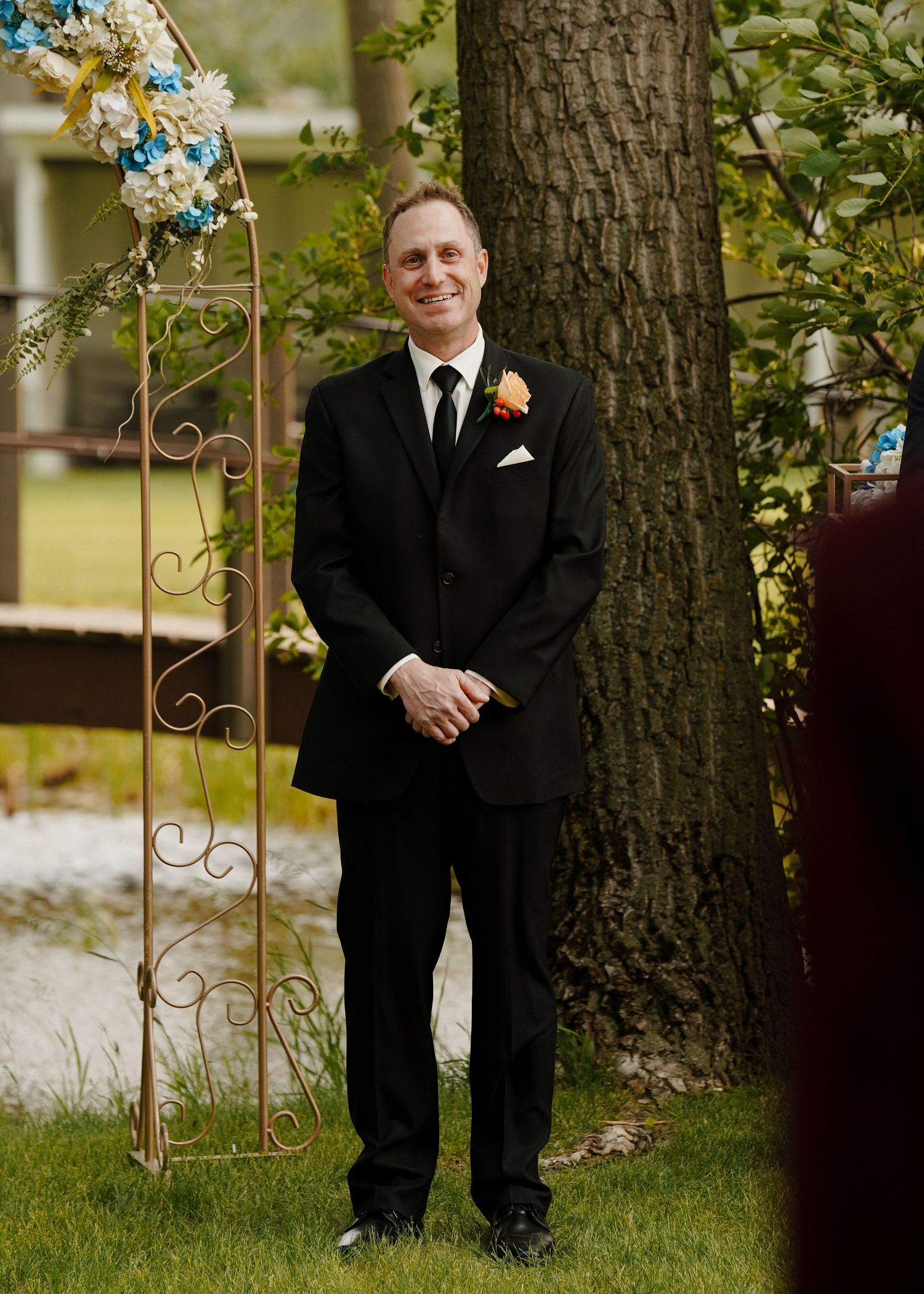 Groom in black suit with orange boutonniere stands smiling beside decorative wedding arch outdoors.