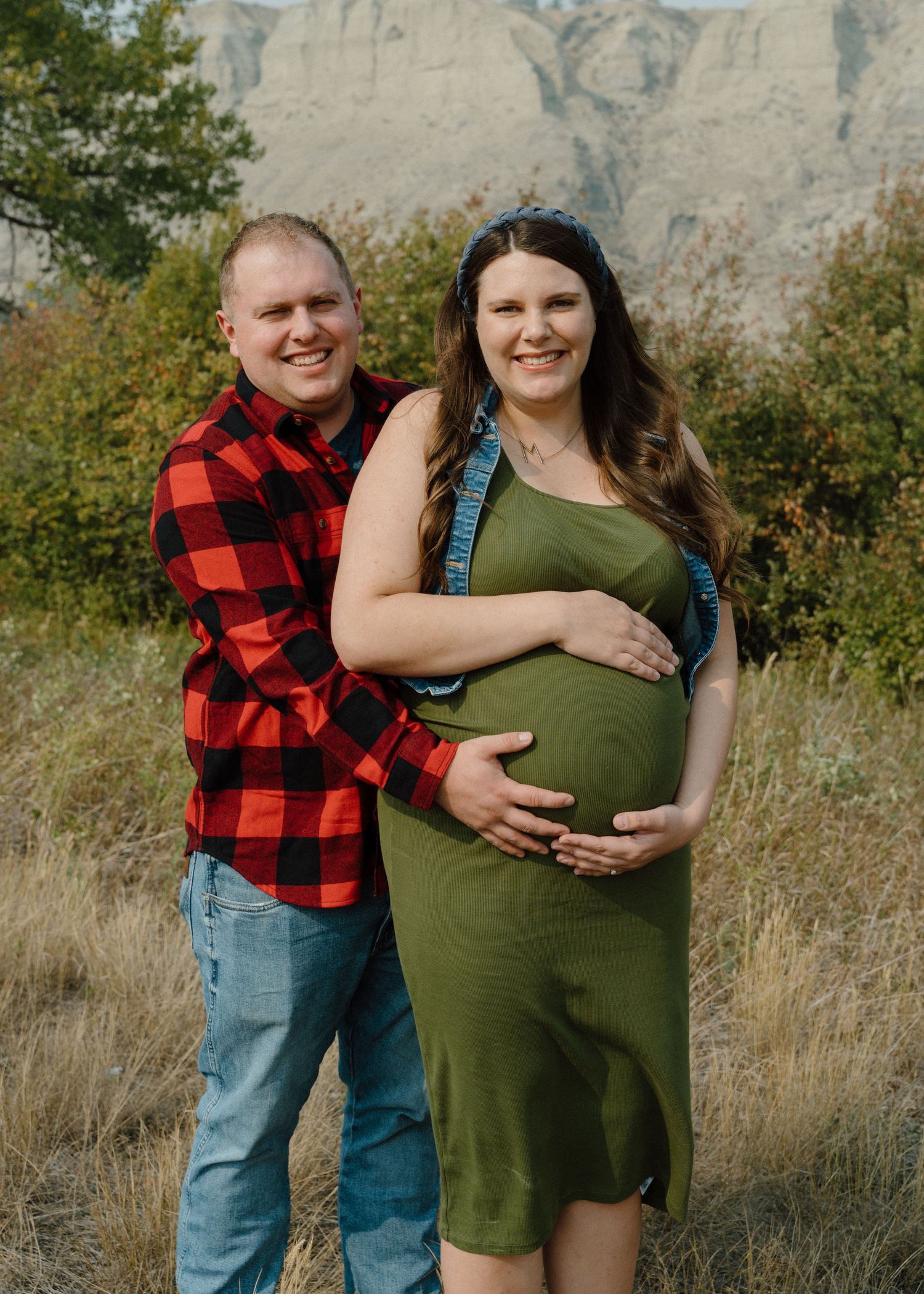 Smiling couple posing outdoors for maternity photos in a field with mountains, woman in green dress showing baby bump.