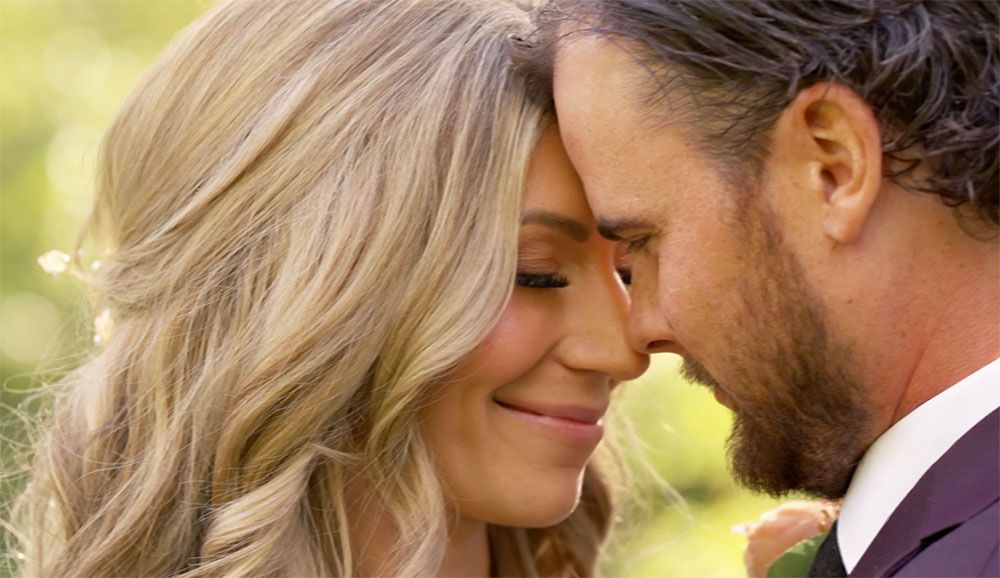 Romantic couple in wedding attire sharing an intimate moment outdoors, touching foreheads and smiling.