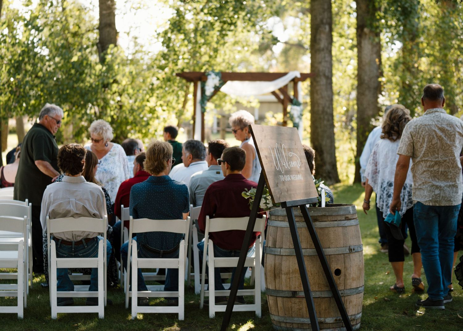 Wedding guests seated in white chairs at outdoor ceremony with rustic wooden barrel and signage.