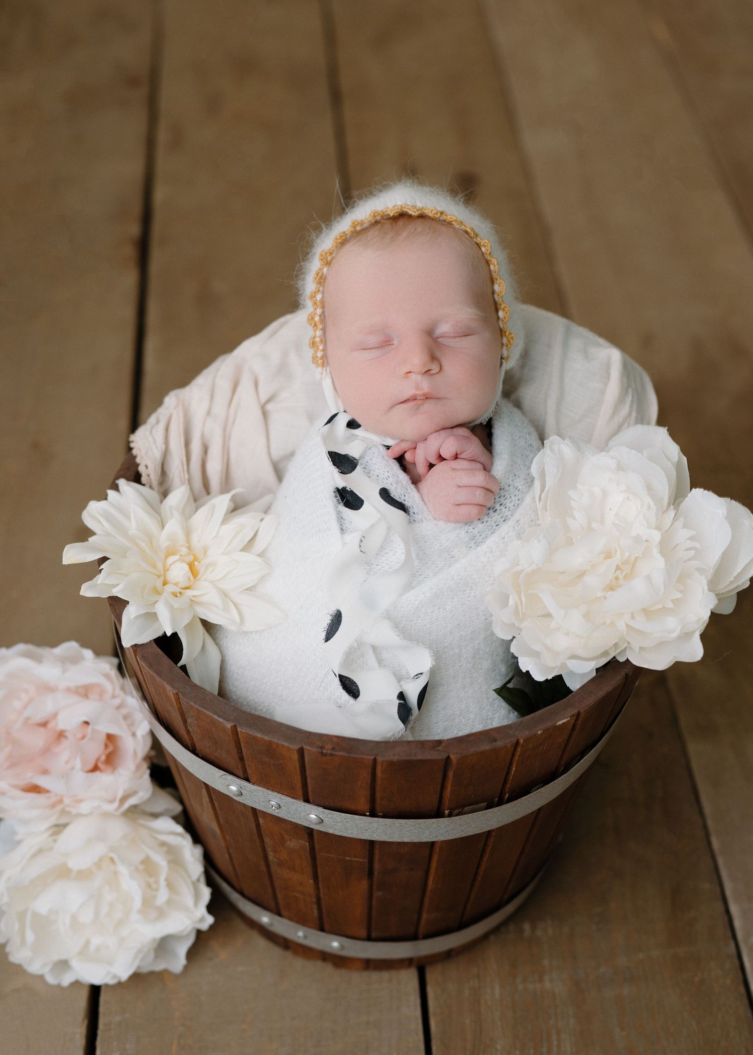 Sleeping newborn in yellow bonnet nestled in wooden bucket surrounded by white peony flowers.