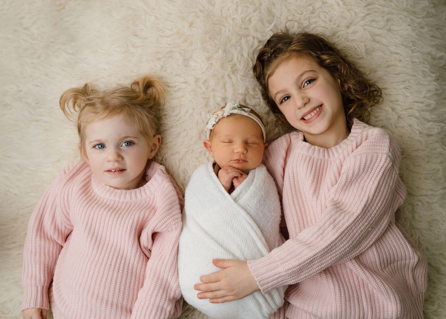 Two older sisters in matching pink sweaters cuddle newborn baby wrapped in white on a fur rug.