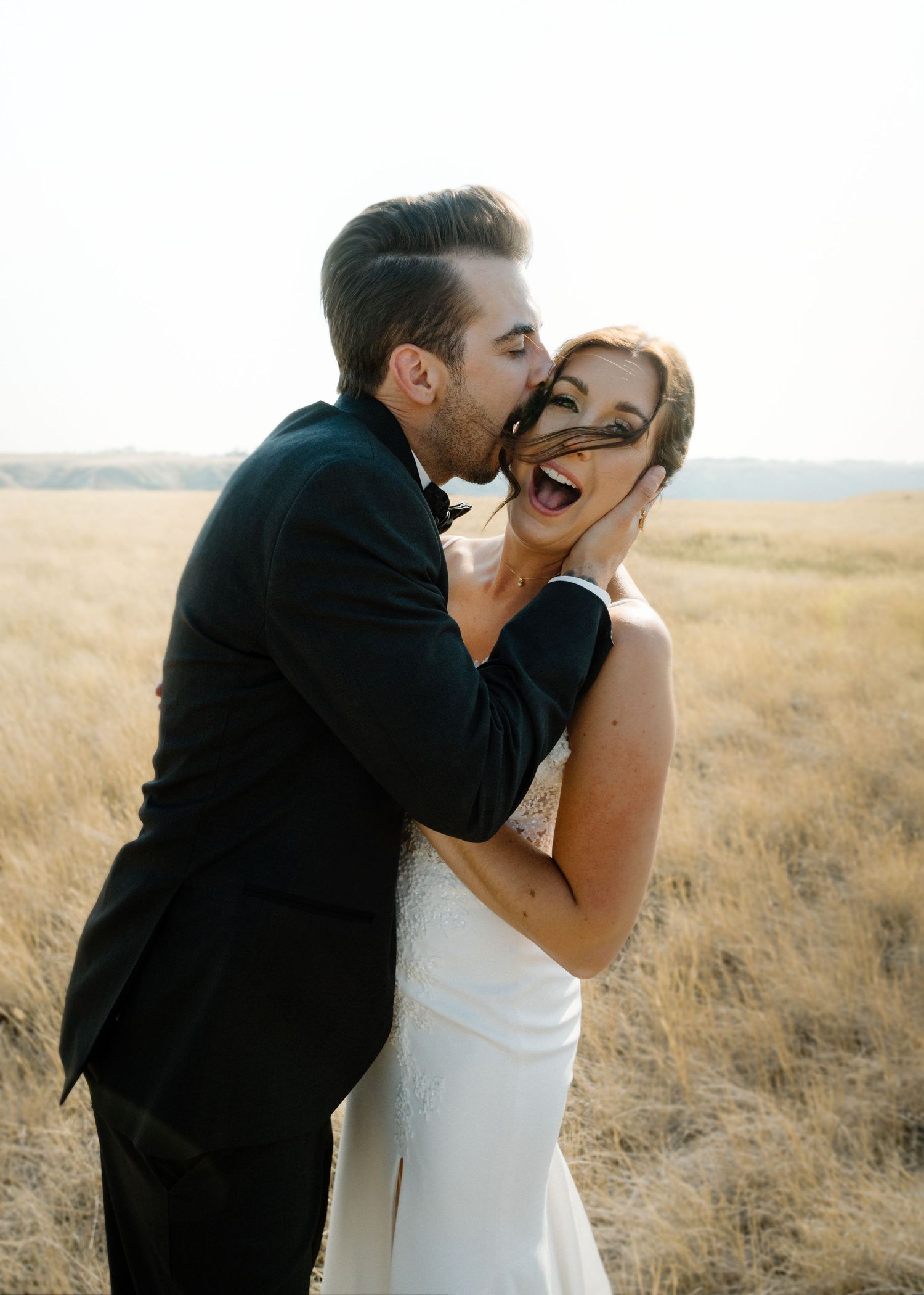 Groom playfully kisses brides cheek as she laughs in a golden prairie field at sunset.
