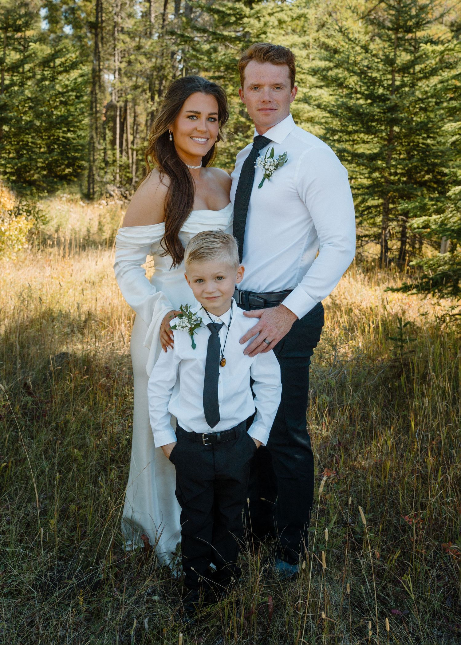 Bride and groom pose with young boy in matching wedding attire in a sunlit forest meadow.