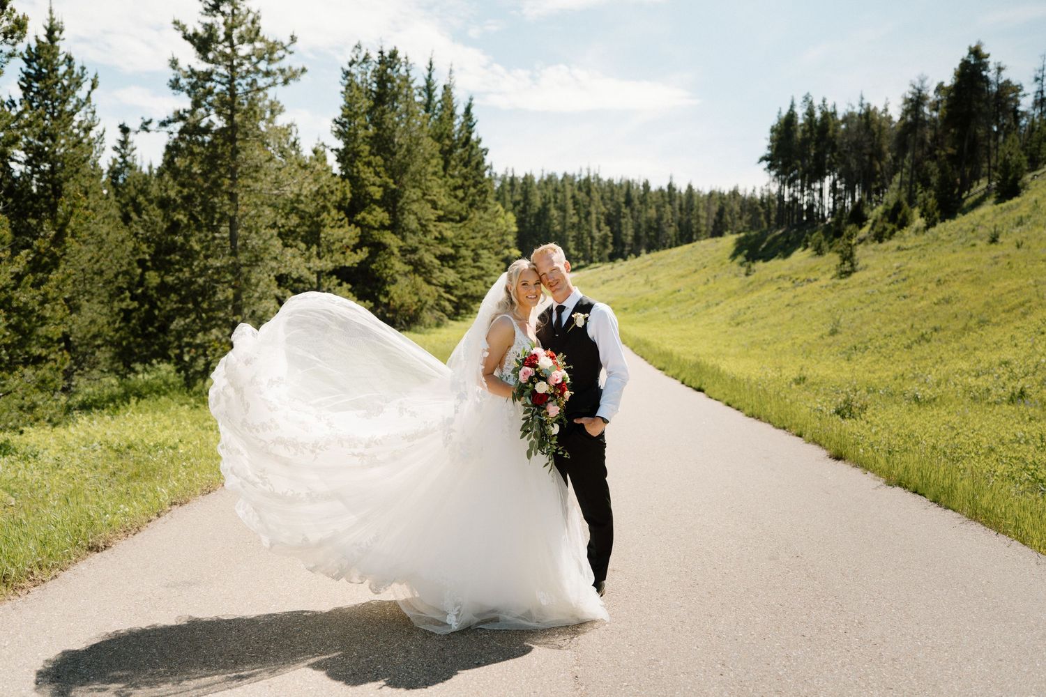 Bride and groom kissing on a scenic mountain road, wedding dress flowing in the wind, surrounded by lush green trees.