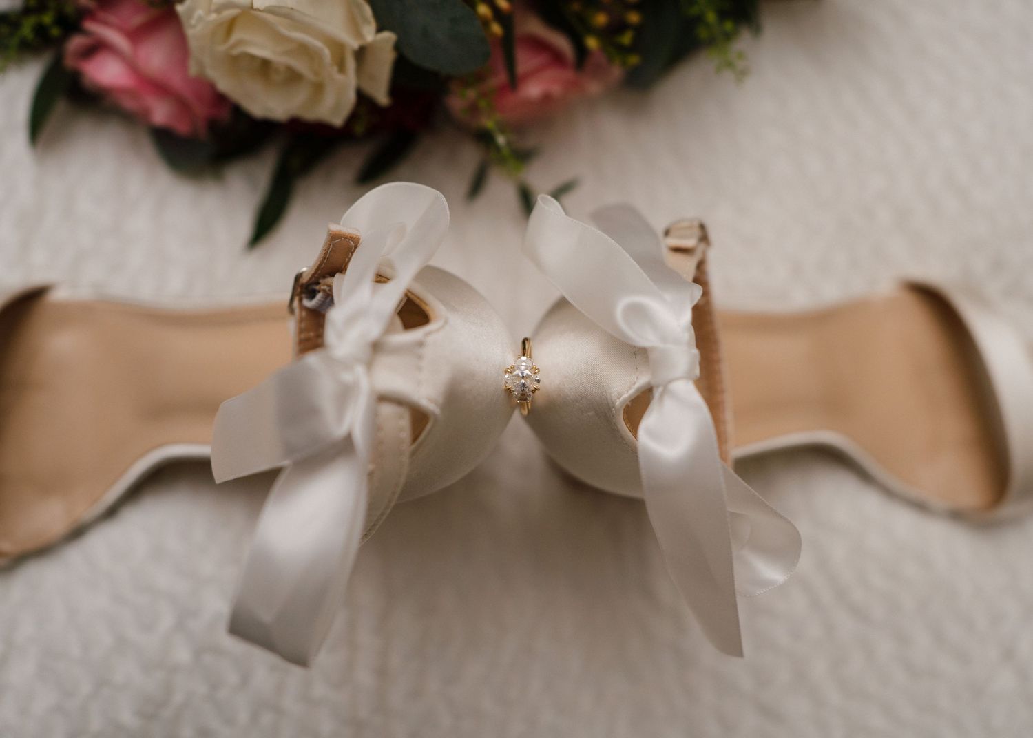 Elegant bridal heels with white satin ribbon bows displayed sole-up near pink and white wedding flowers.