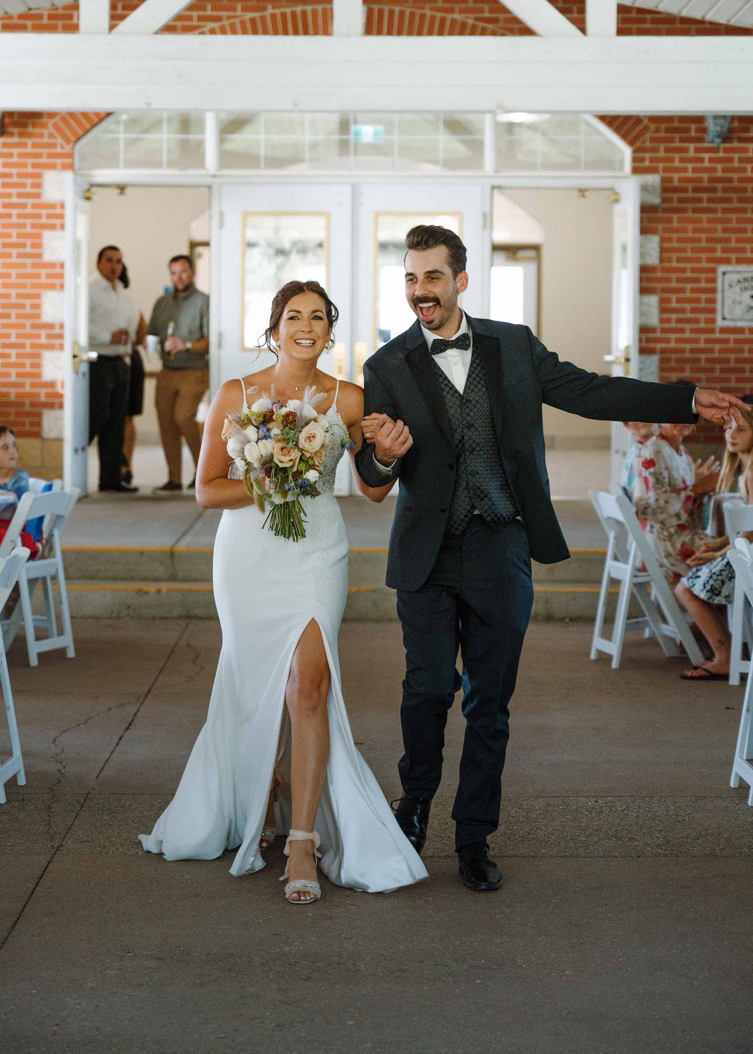 Joyful bride and groom walk down the aisle after wedding ceremony at a brick venue, bride holding a bouquet.
